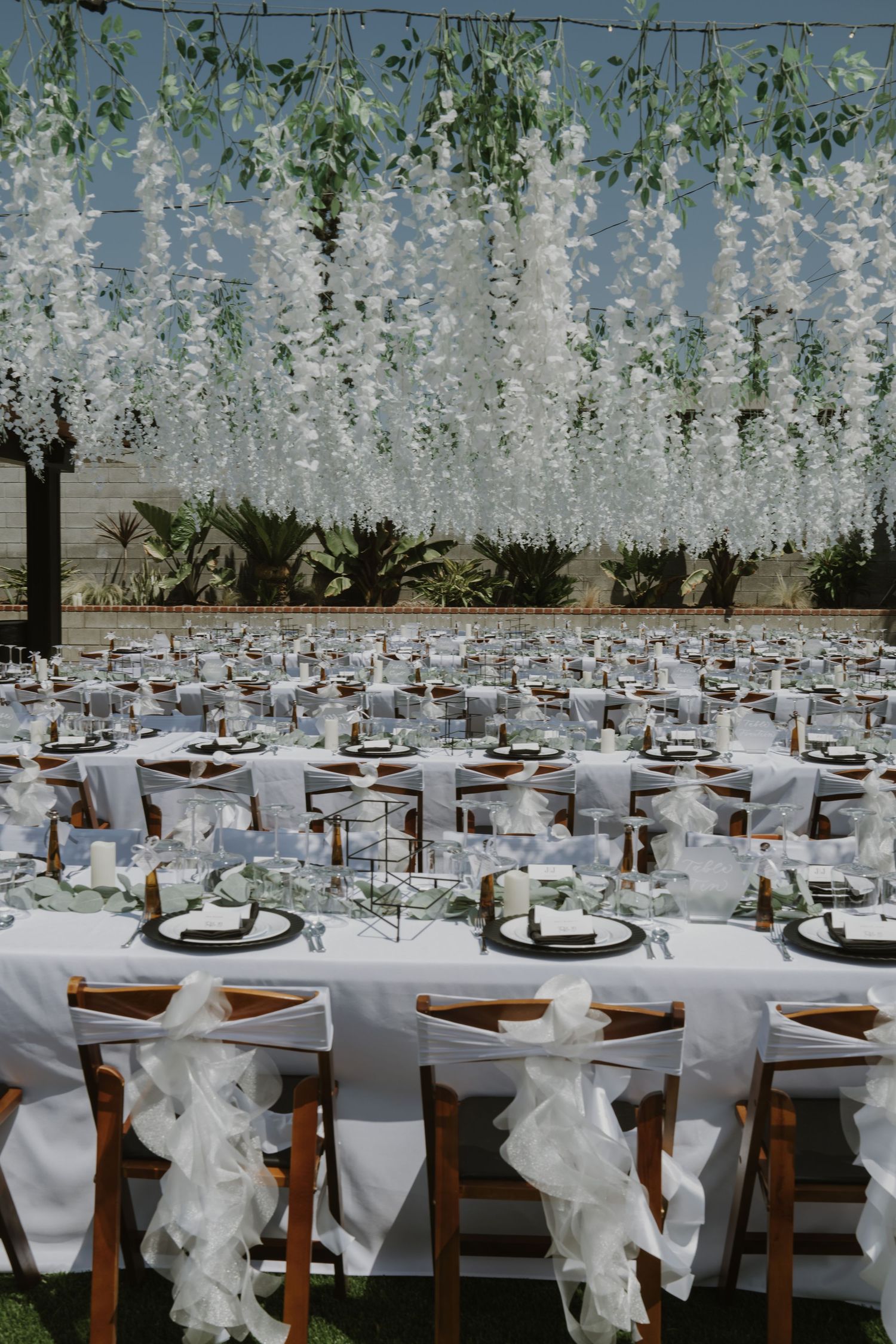 Elegant outdoor wedding reception with white floral ceiling installation, draped chairs, and formally set tables.