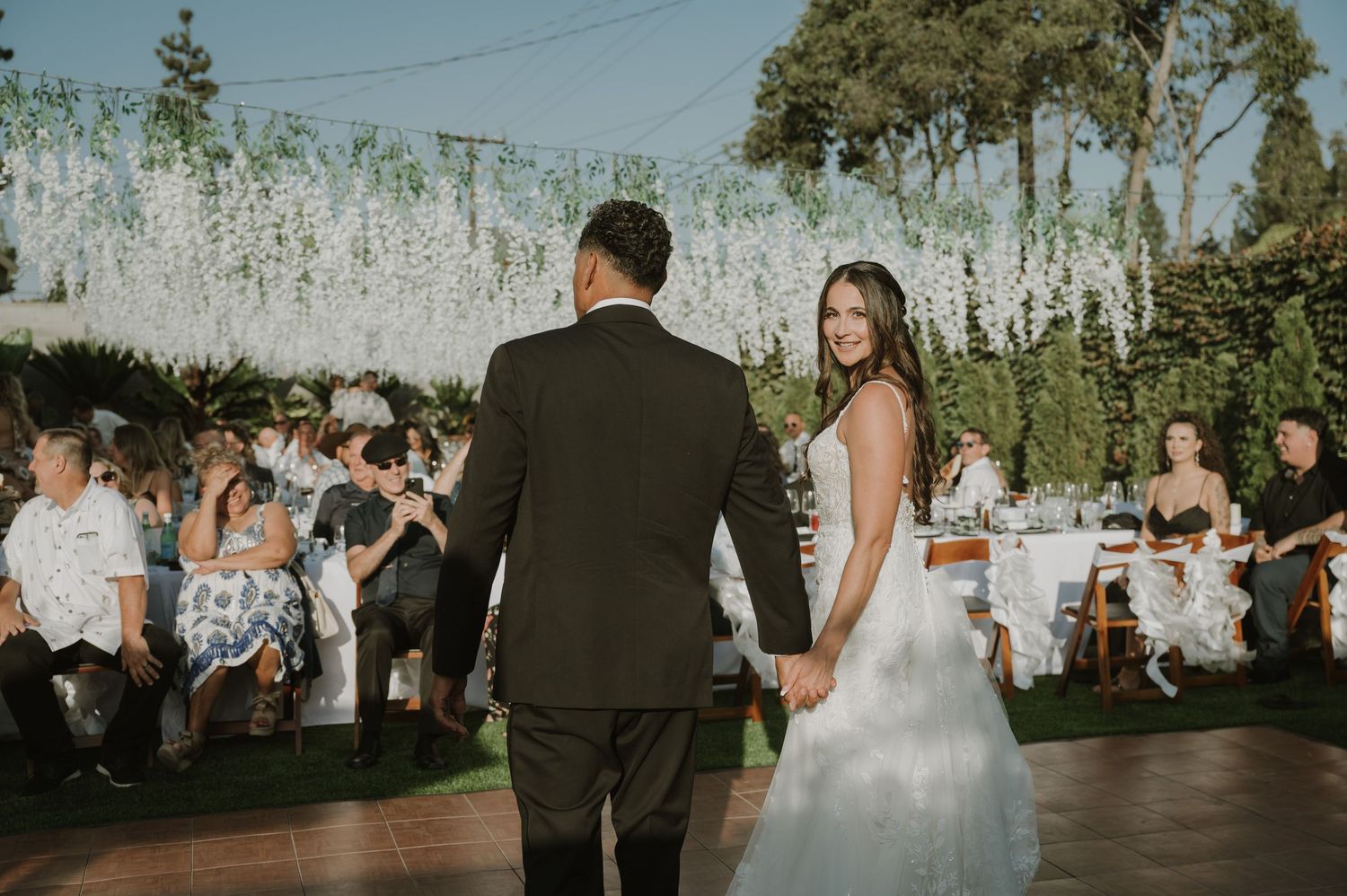 Bride and groom share first dance outdoors at elegant wedding reception with white floral ceiling decor and seated guests.