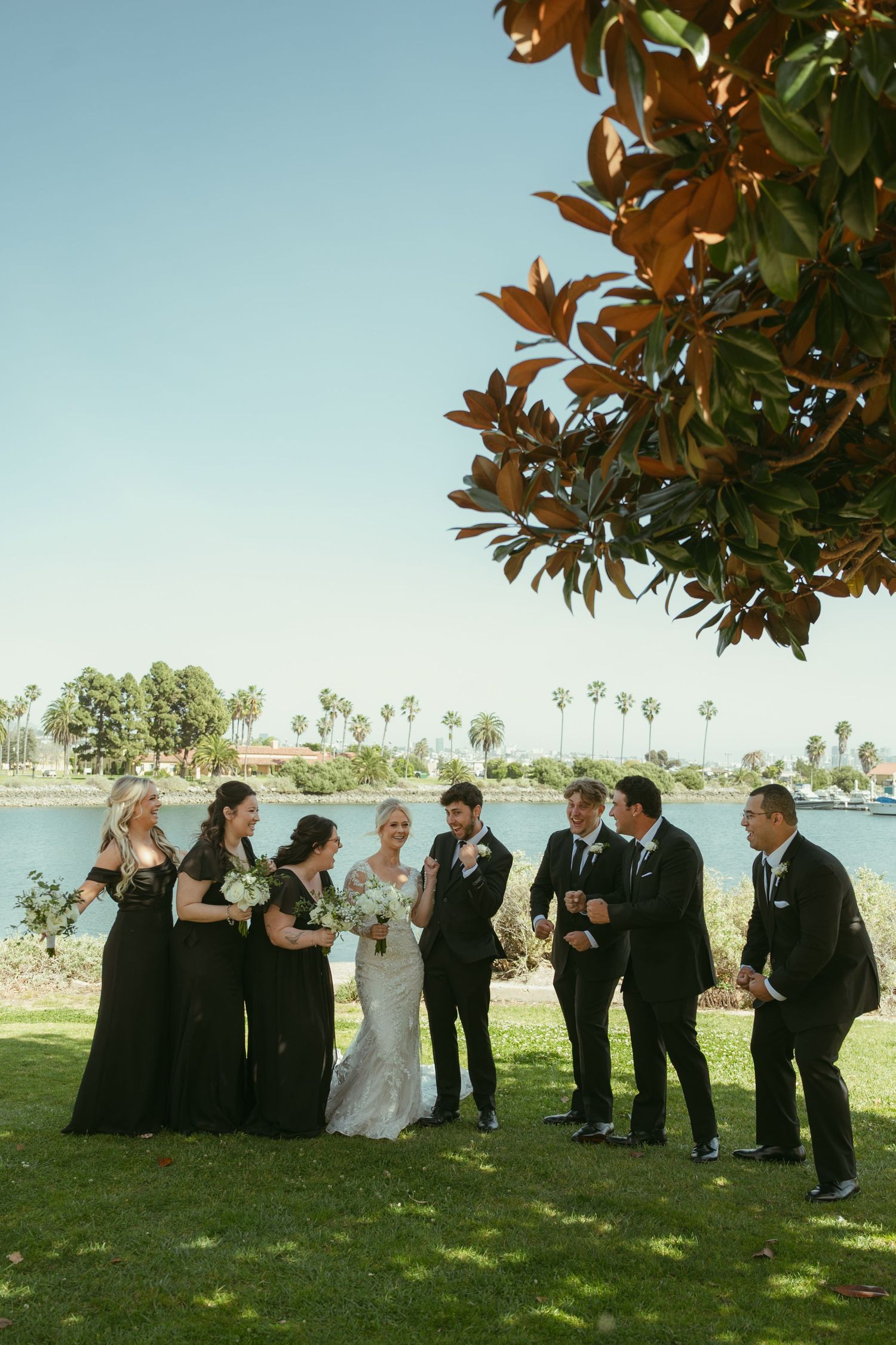Wedding party poses outdoors near a lake with palm trees, bridesmaids in black dresses and groomsmen in suits.