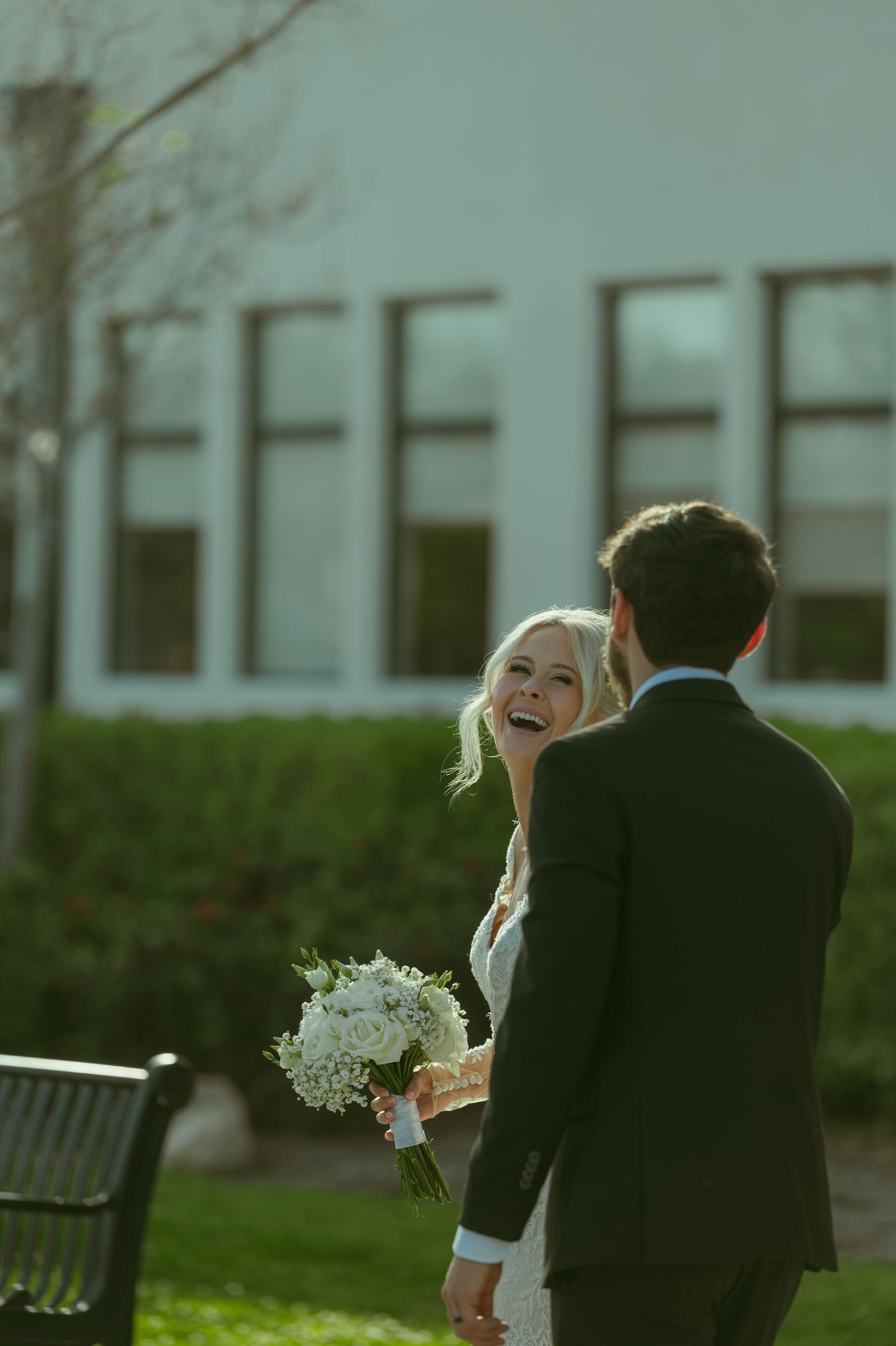 Smiling bride holding white bouquet laughs with groom in dark suit outdoors near white building on wedding day.