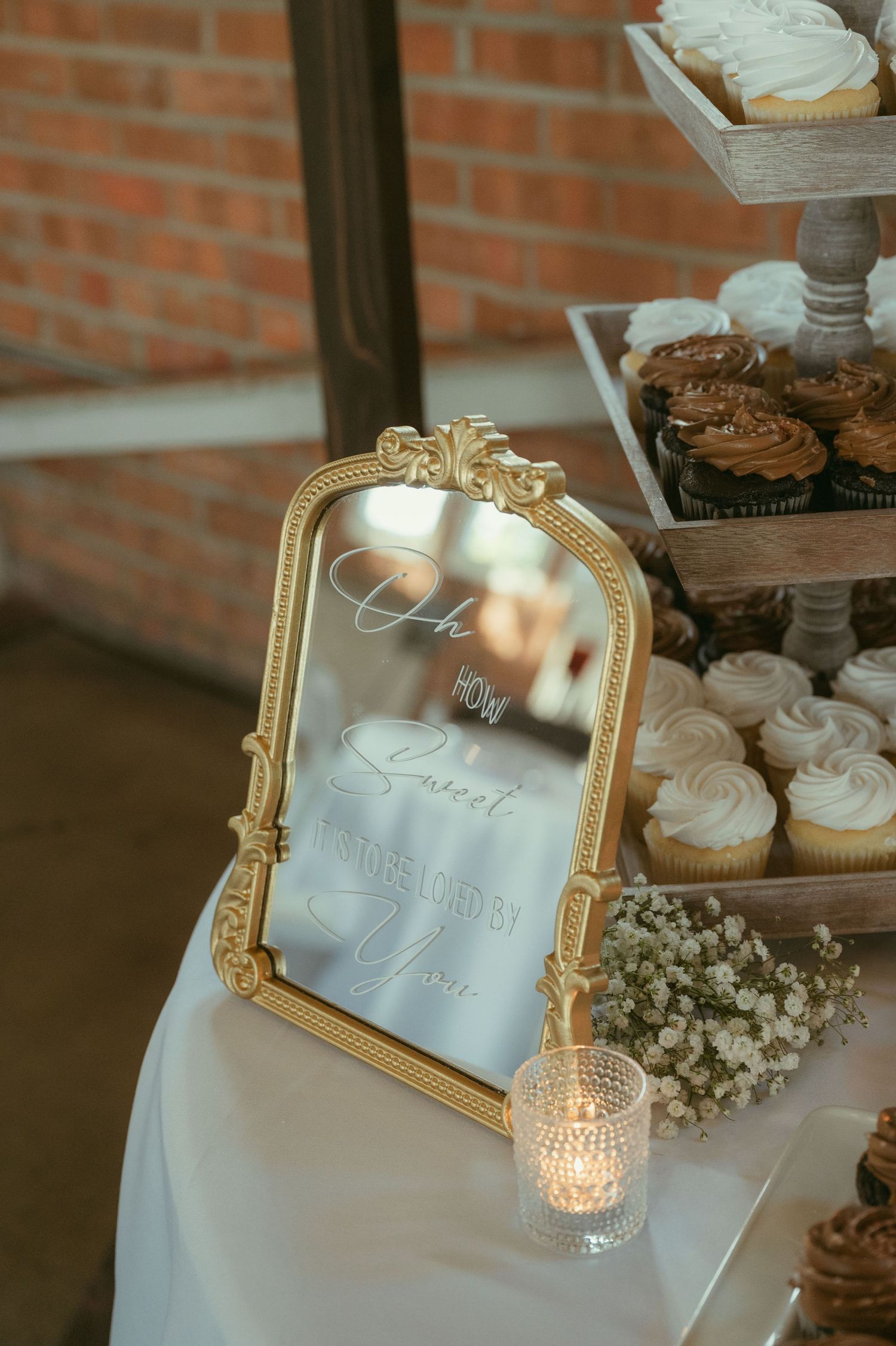Gold ornate mirror sign on a wedding dessert table with cupcakes, baby's breath, and a candle.