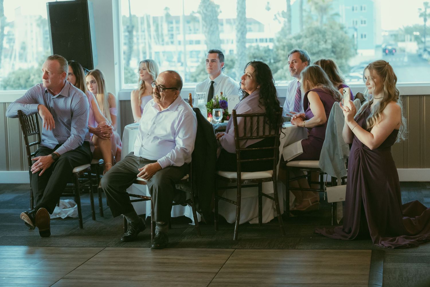 Wedding reception guests seated and standing in a bright venue with palm tree views outside.