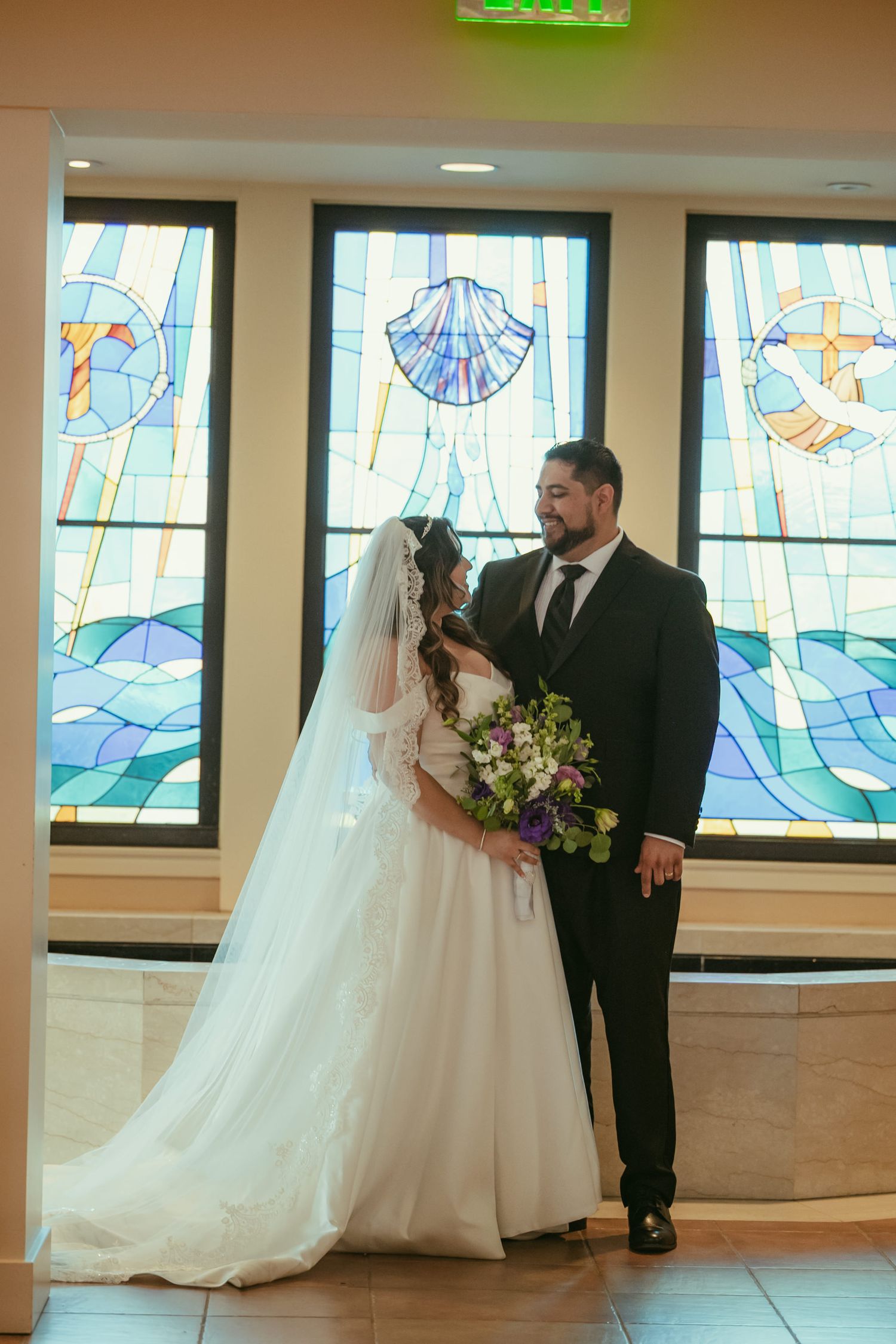 Bride and groom stand before colorful stained glass windows in a church, bride holding a purple and green bouquet.