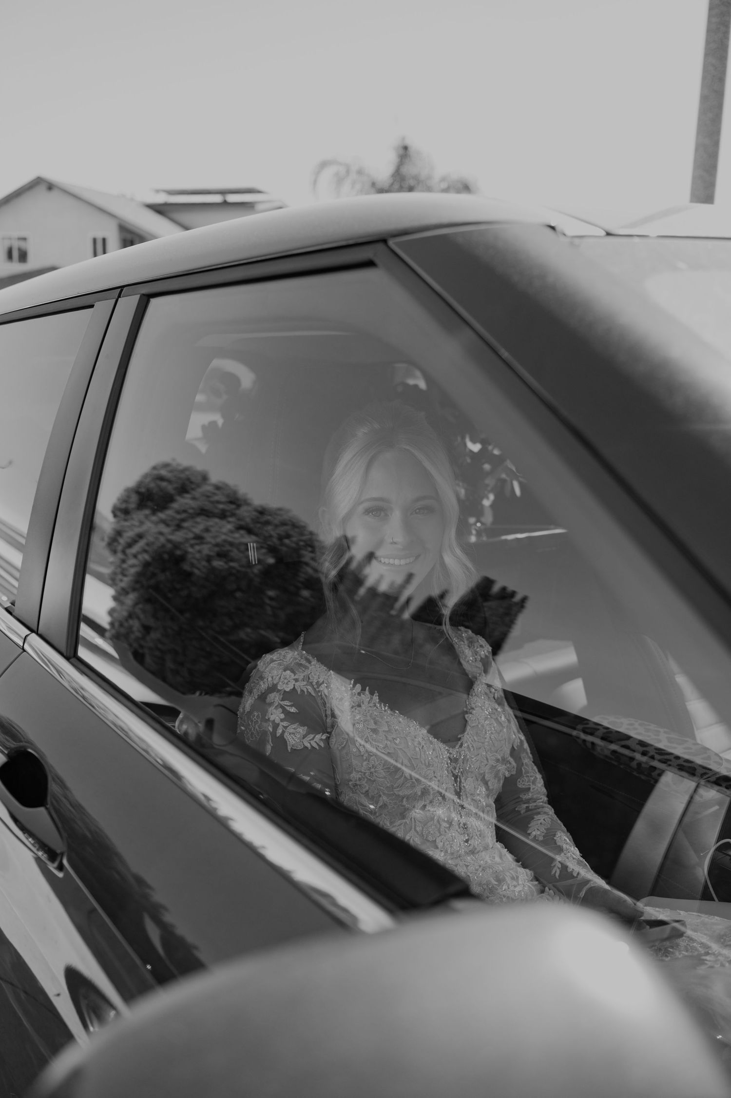 Smiling bride in lace dress sitting in a car with a dark curly-haired dog on her wedding day.