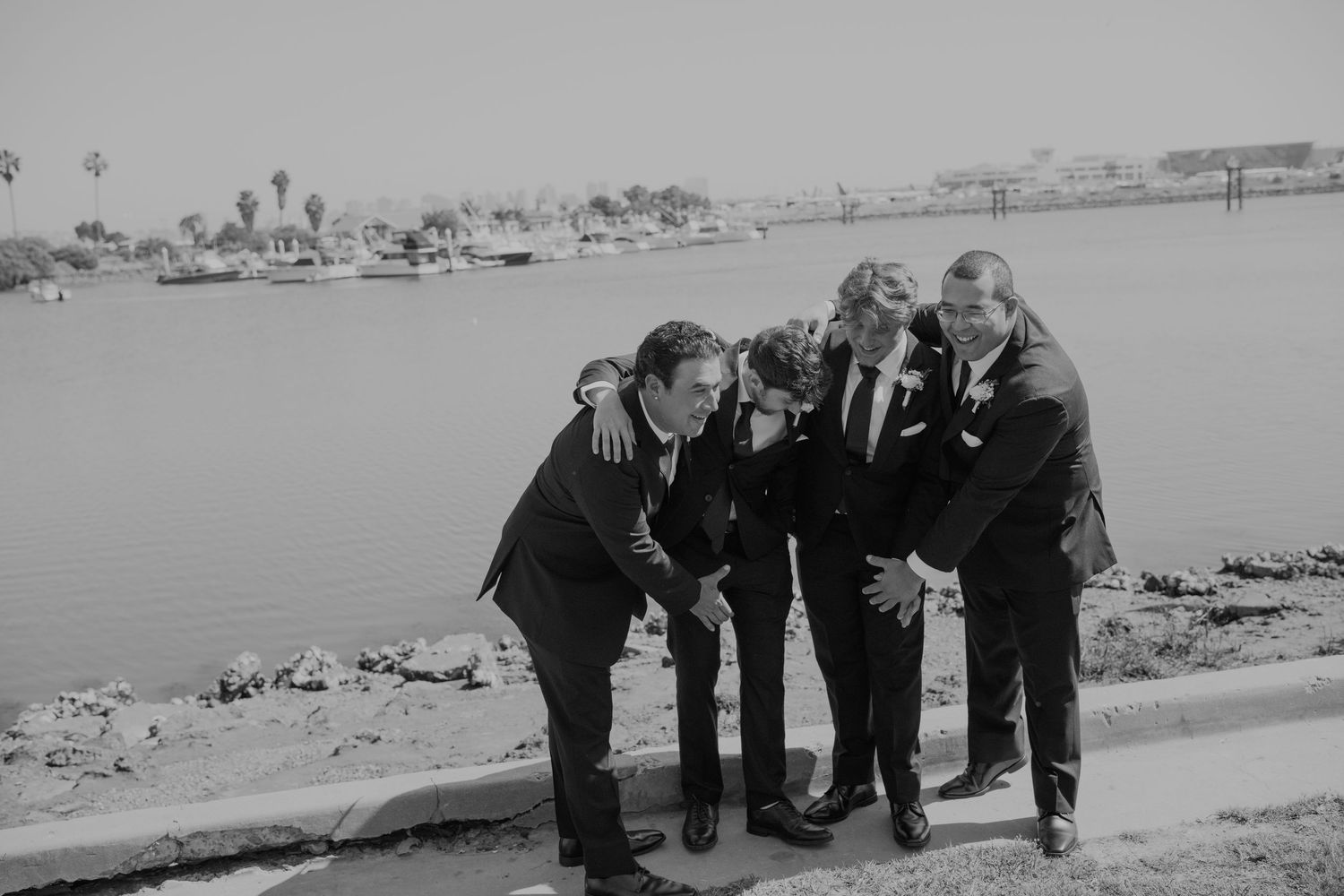 Black and white photo of four groomsmen in suits laughing together near a waterfront.