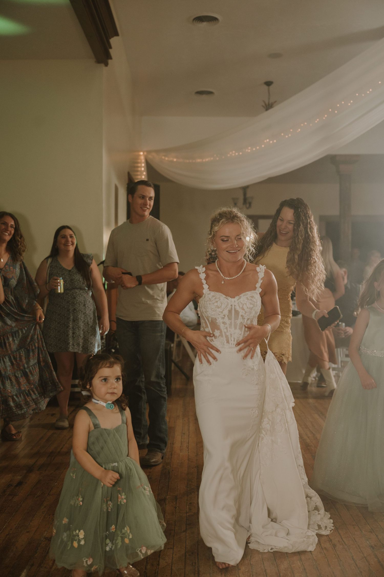 Wedding celebration showing guests dancing together in a rustic venue with white draping and string lights.