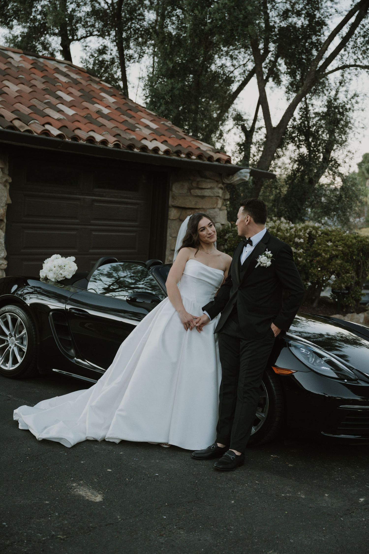 A couple poses in formal attire next to luxury cars outside a Spanish-style building.