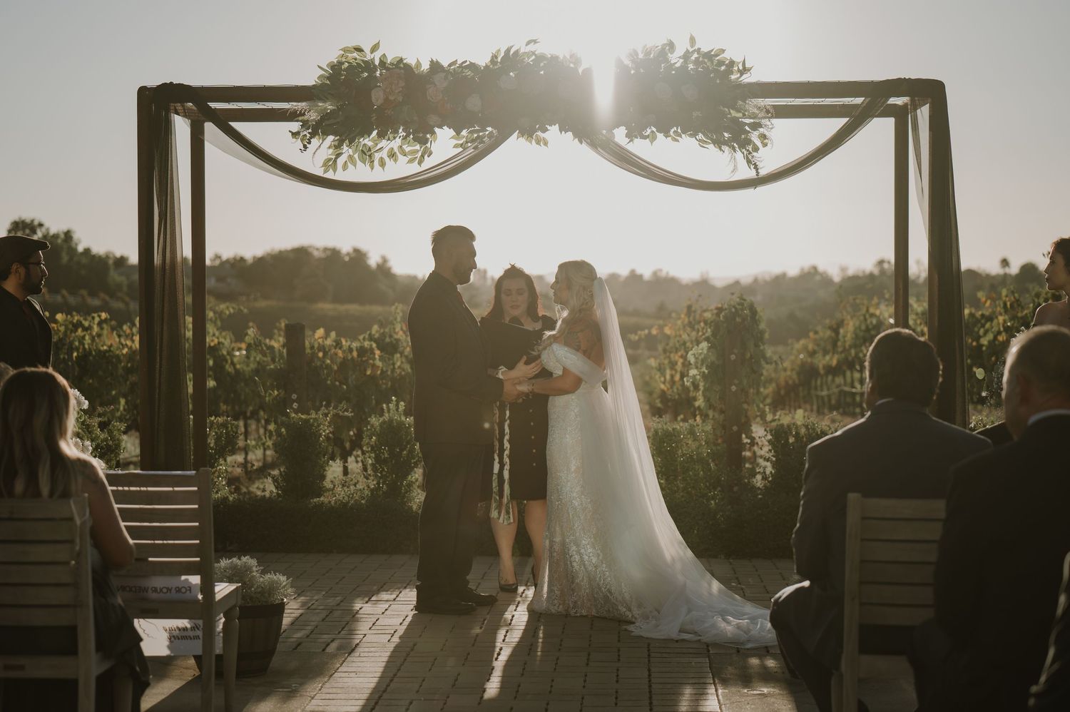 Sunset wedding ceremony silhouette under decorated wooden arch with guests seated.