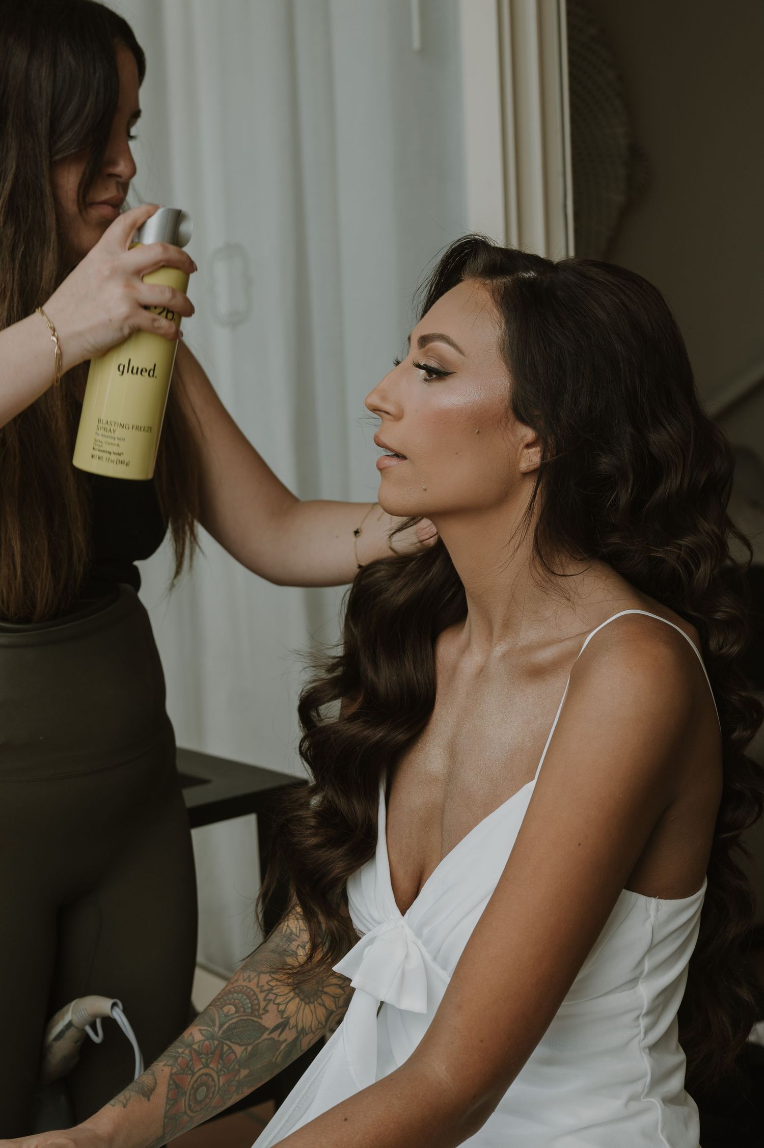 Woman in white dress getting makeup applied in moody lighting.