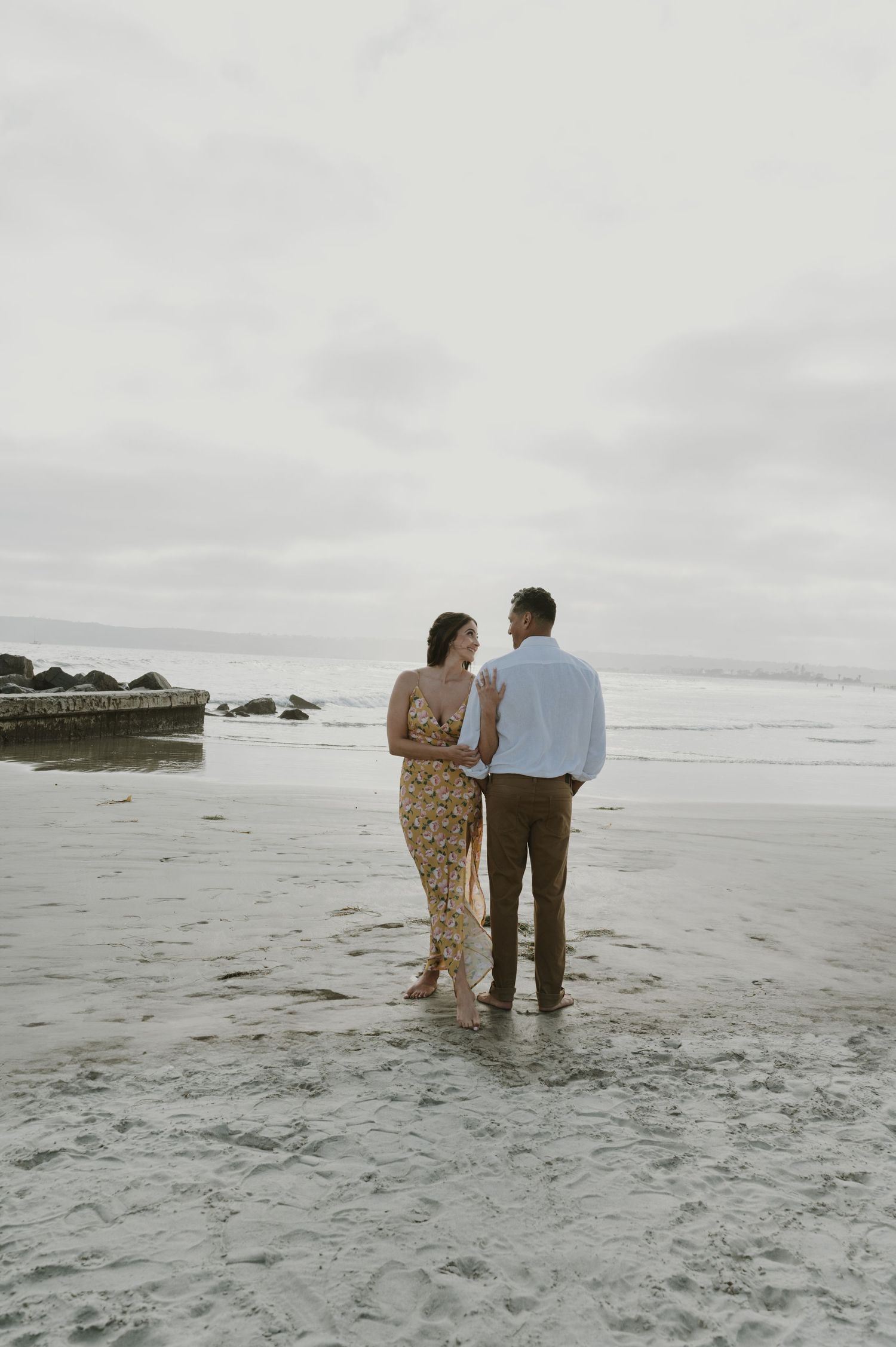 Couple walking together on a misty beach shore with waves and rocks in the background.