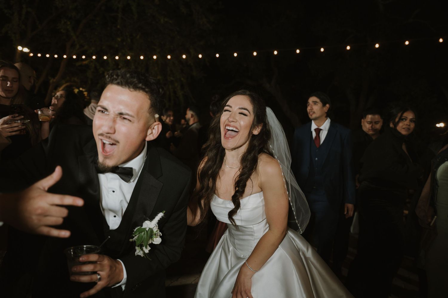 Guests celebrating at an outdoor wedding reception with string lights overhead.