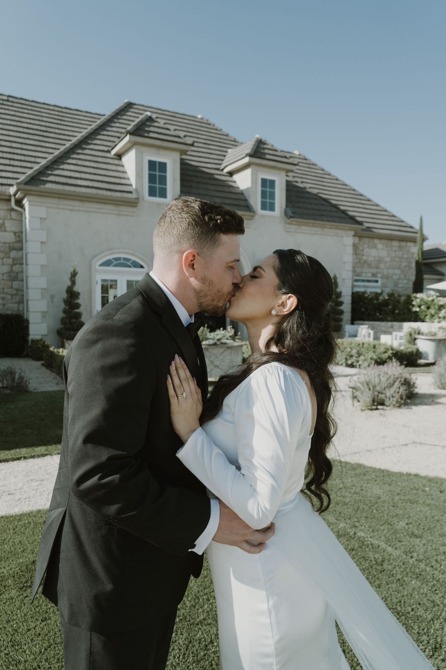 A romantic outdoor wedding photoshoot showing a couple sharing tender kisses in front of a stone house.