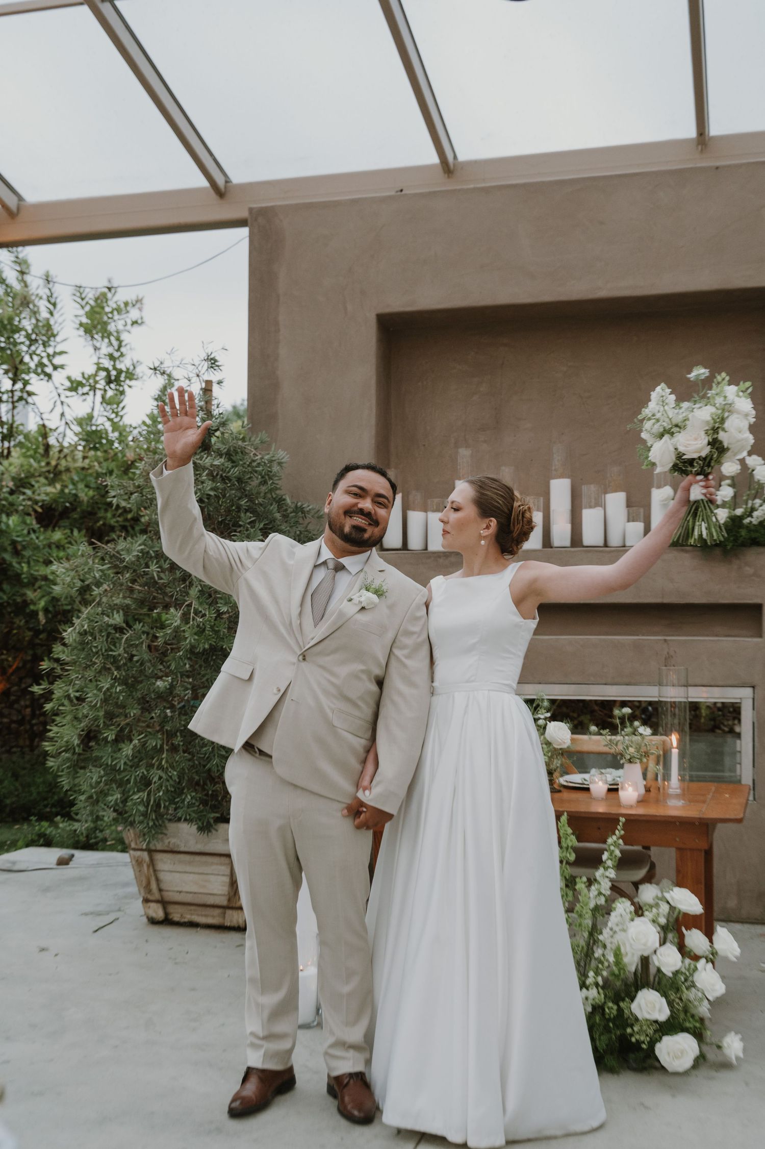 Newlywed couple celebrating at outdoor wedding ceremony, groom in beige suit, bride in white gown holding bouquet.