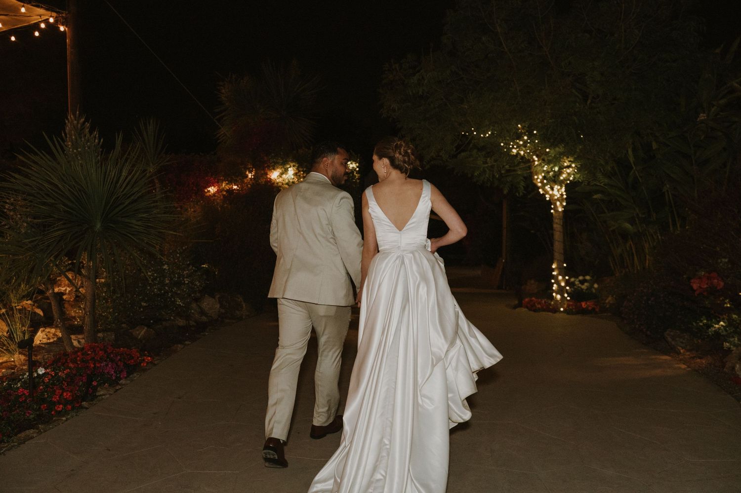 Bride and groom walking hand in hand through illuminated garden path at night wedding.