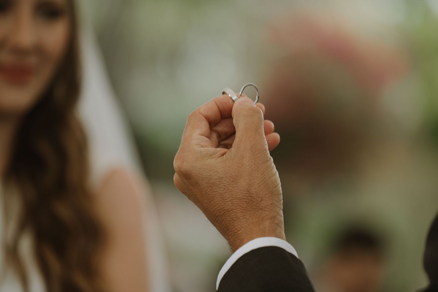 Groom holding wedding rings during outdoor ceremony with bride in white veil smiling in background.