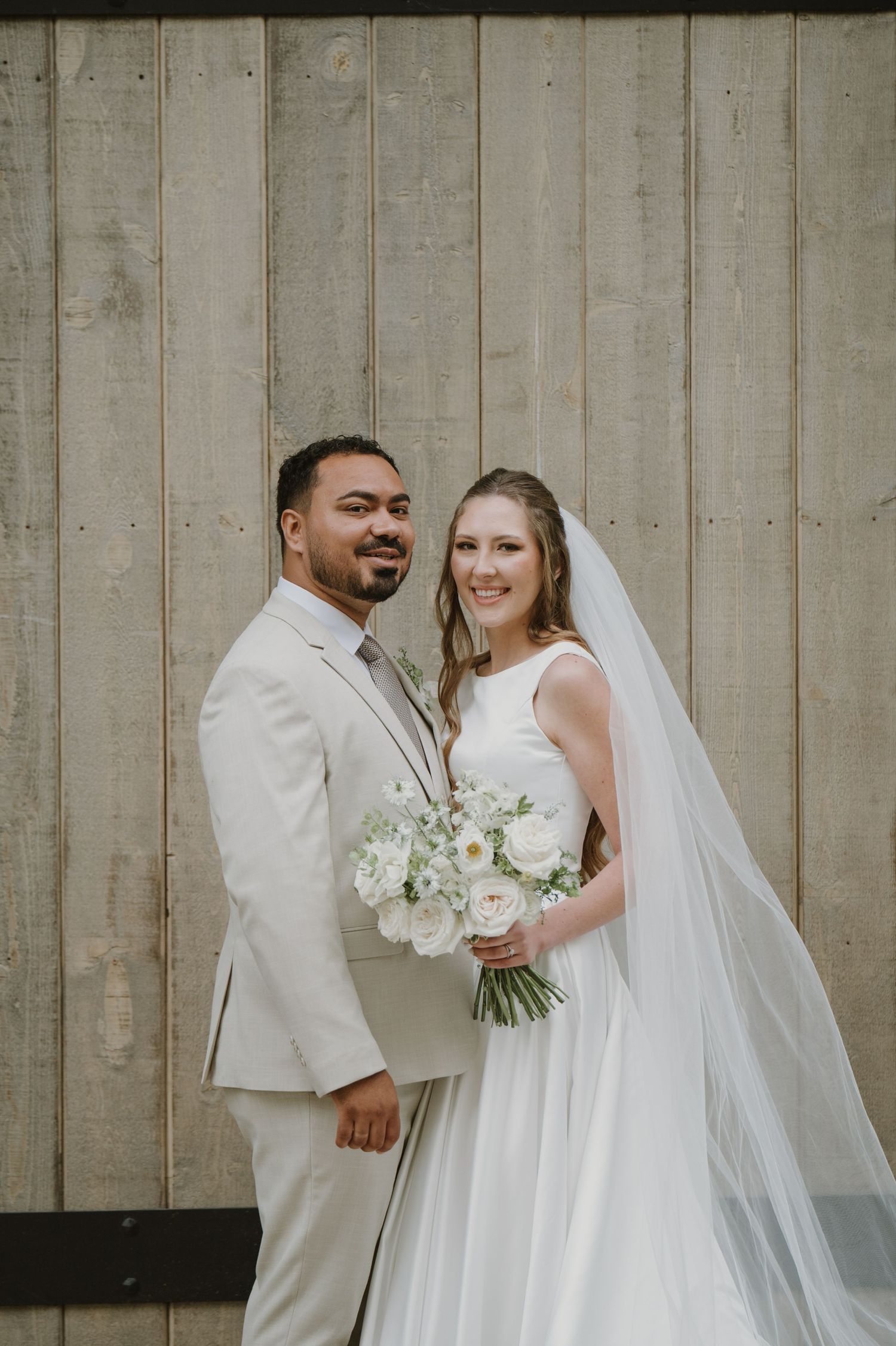 Smiling newlyweds pose together, bride holding white floral bouquet with veil, groom in beige suit against wood wall.