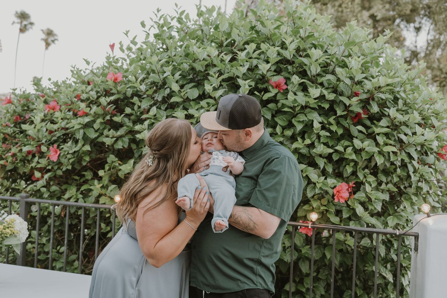 Young couple kissing their newborn baby outdoors in front of a lush green bush with pink flowers.