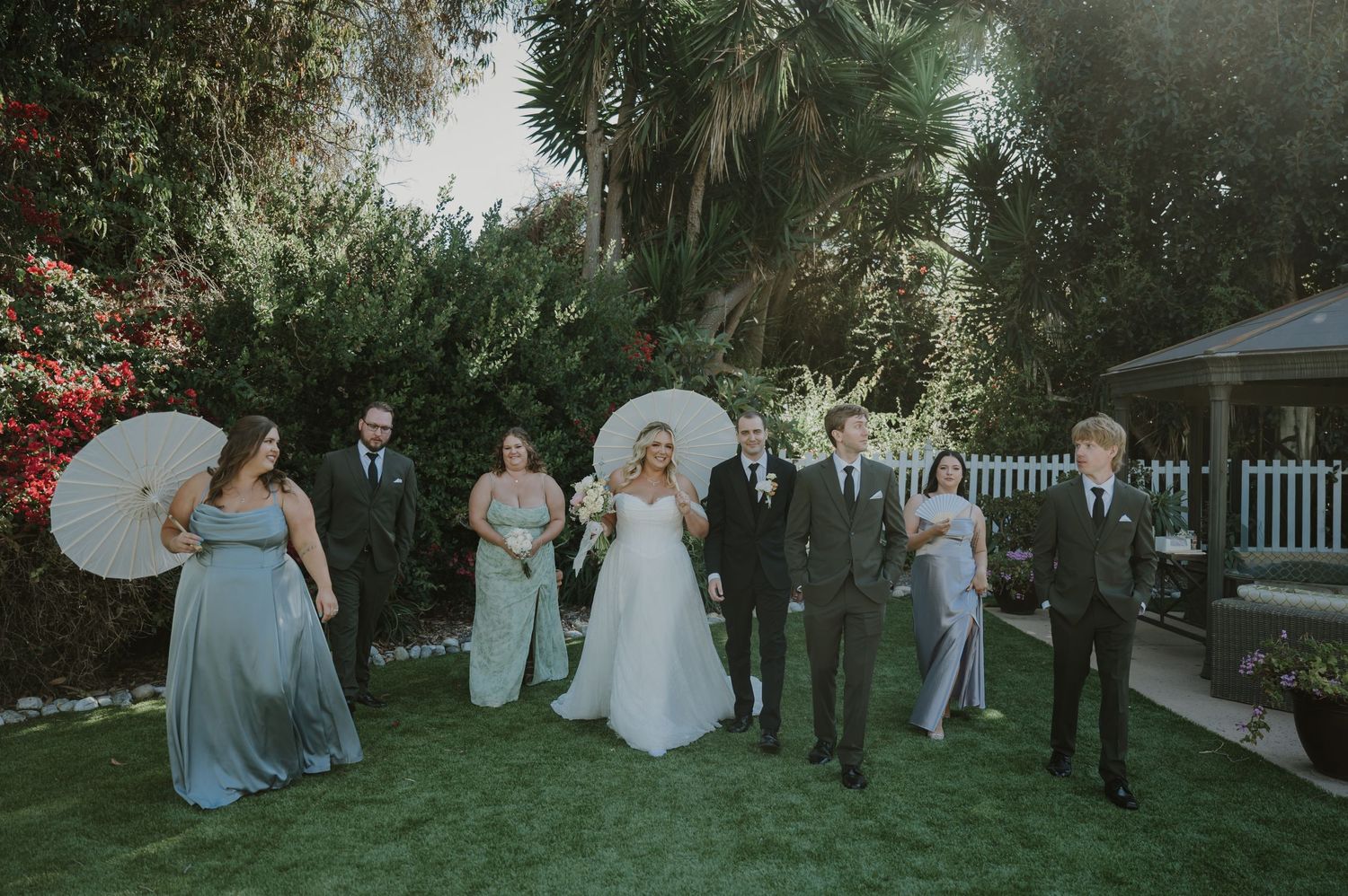 Wedding party poses on green lawn with bride, groom, bridesmaids in sage, and groomsmen holding parasols.