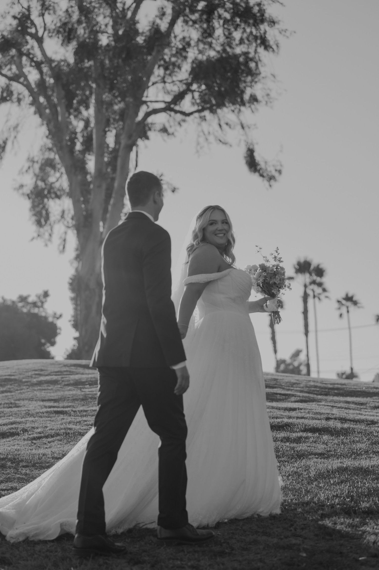 Black and white photo of a bride in a flowing gown holding a bouquet, smiling at the groom outdoors.