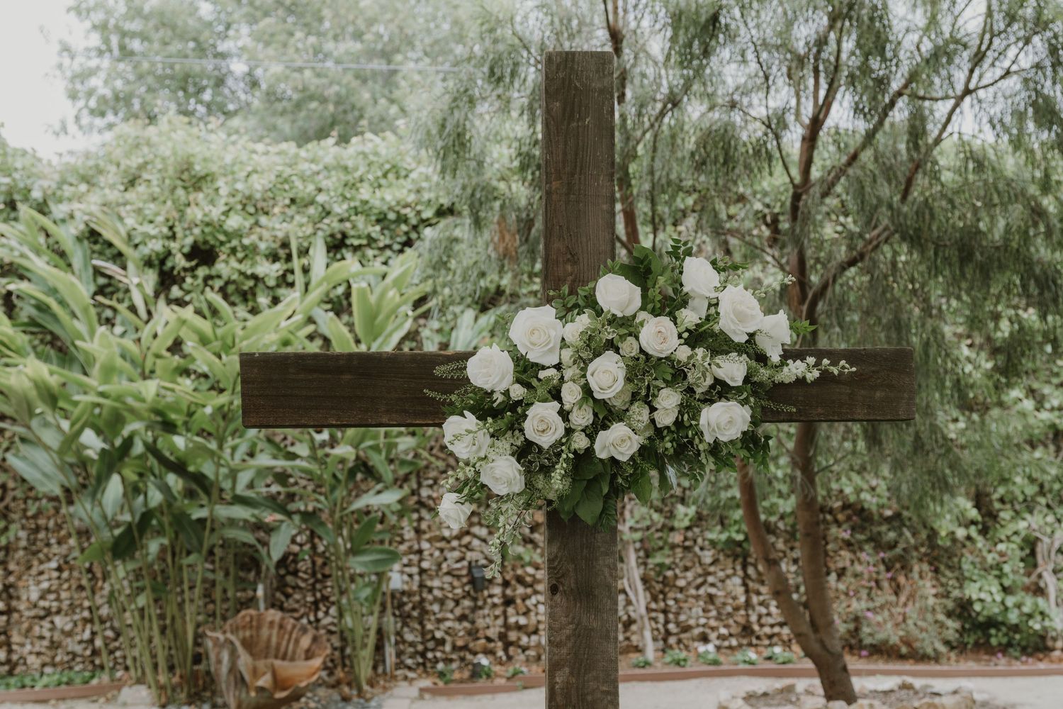 Rustic wooden cross adorned with white roses and greenery floral arrangement at outdoor wedding ceremony.