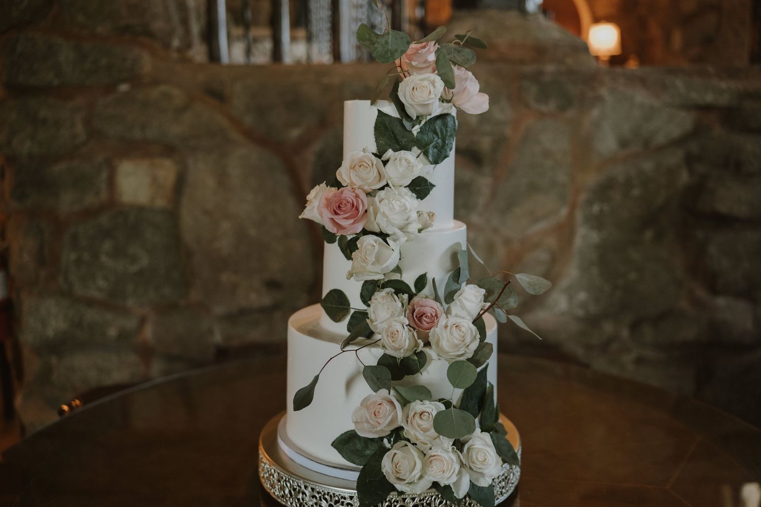 Elegant four-tier white wedding cake adorned with cascading pink and ivory roses and greenery on a decorative silver stand.