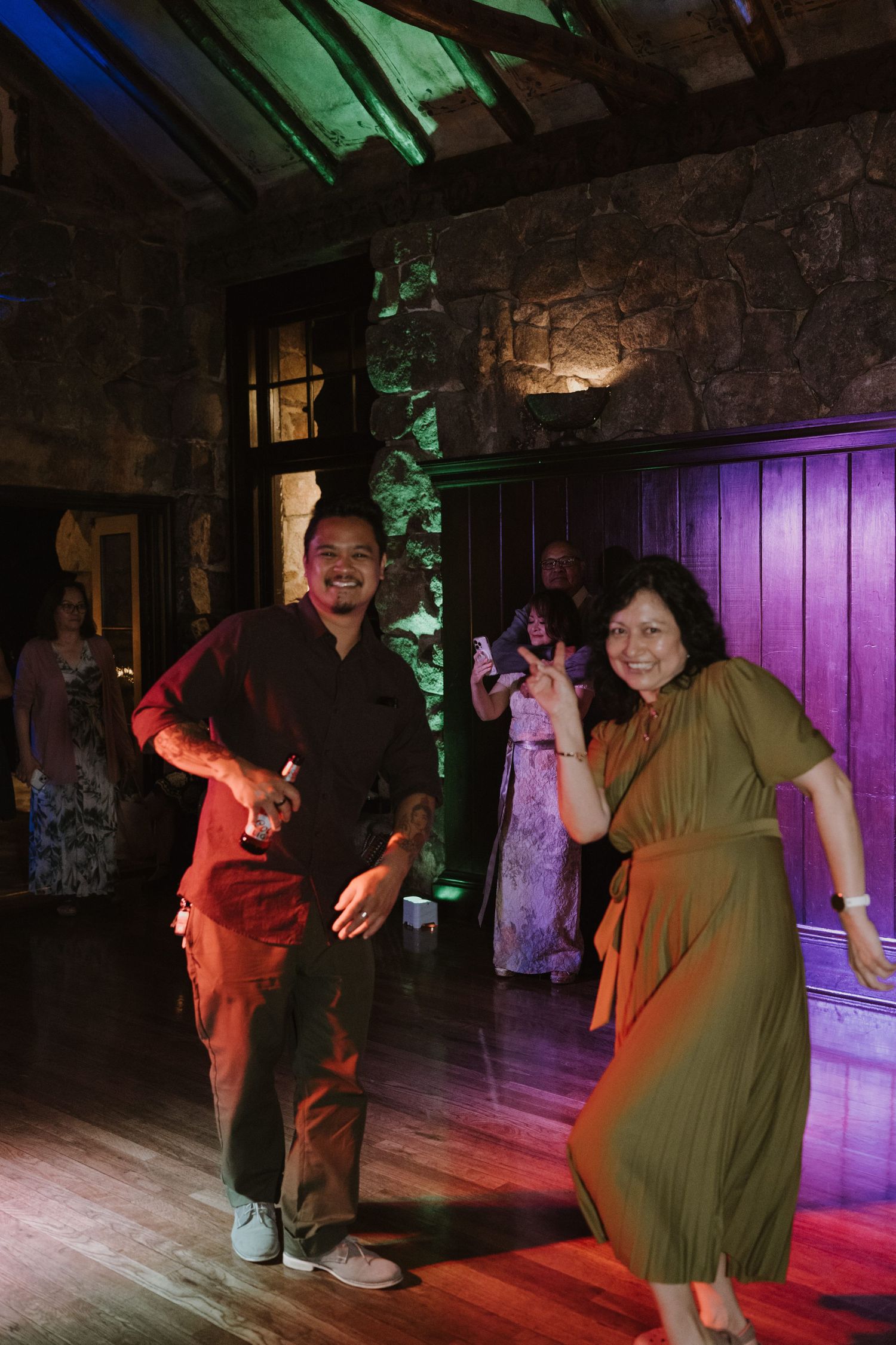 Two guests dancing joyfully on a wooden dance floor at a wedding reception in a rustic stone venue.