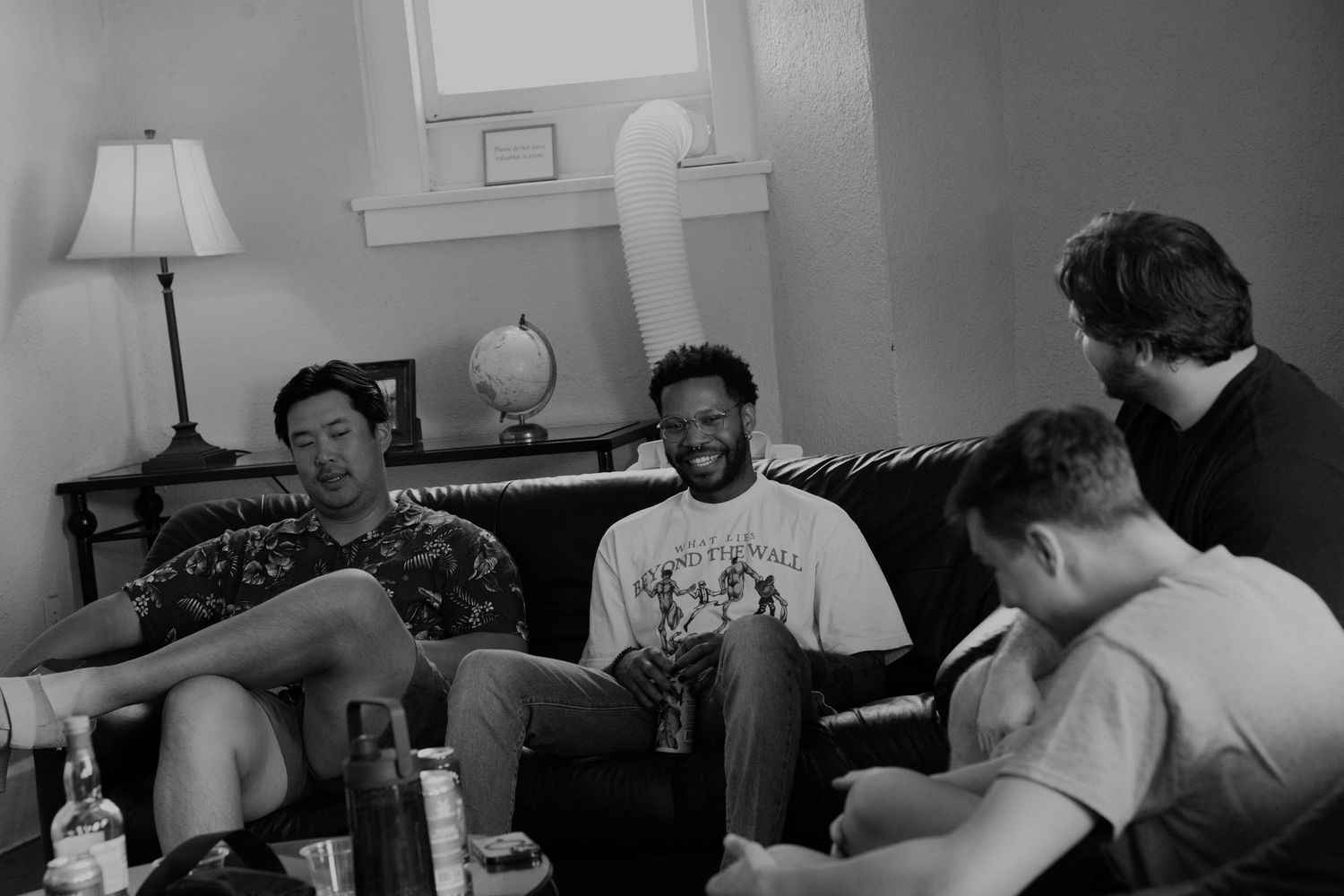 Four young men laughing and socializing together on a couch in a living room, black and white photo.