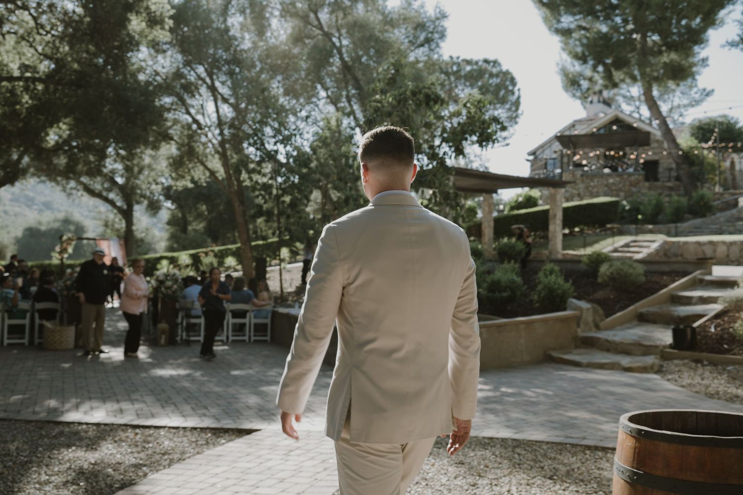 Groom in beige suit walks toward outdoor wedding ceremony venue surrounded by trees and guests.