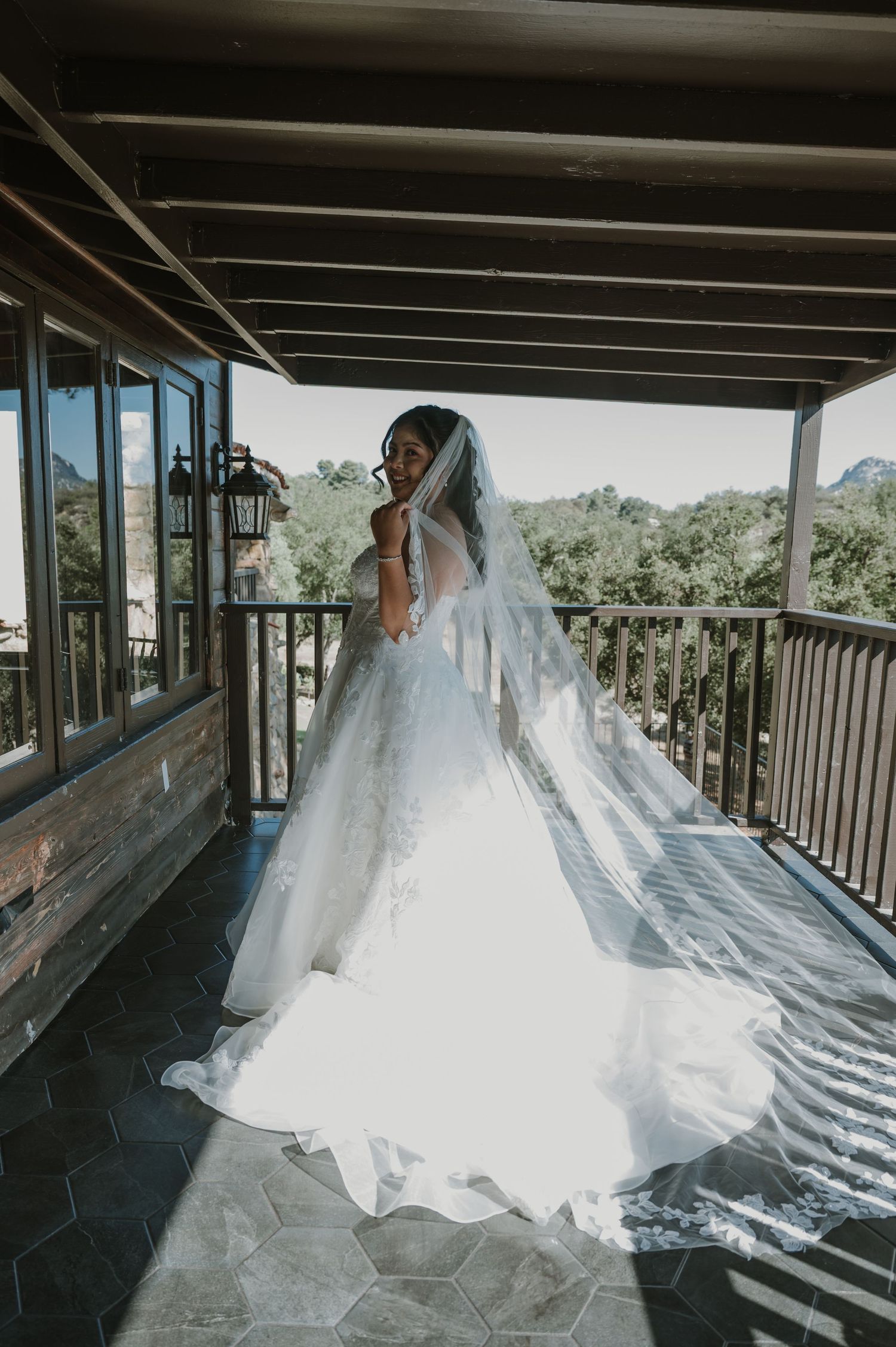 Bride in white ball gown and long veil stands on a rustic balcony surrounded by lush greenery.