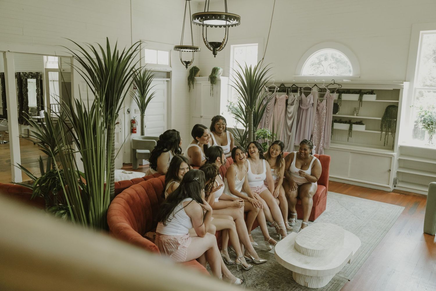 Bridesmaids in white loungewear gathered on red sofa in bright bridal suite before wedding ceremony.
