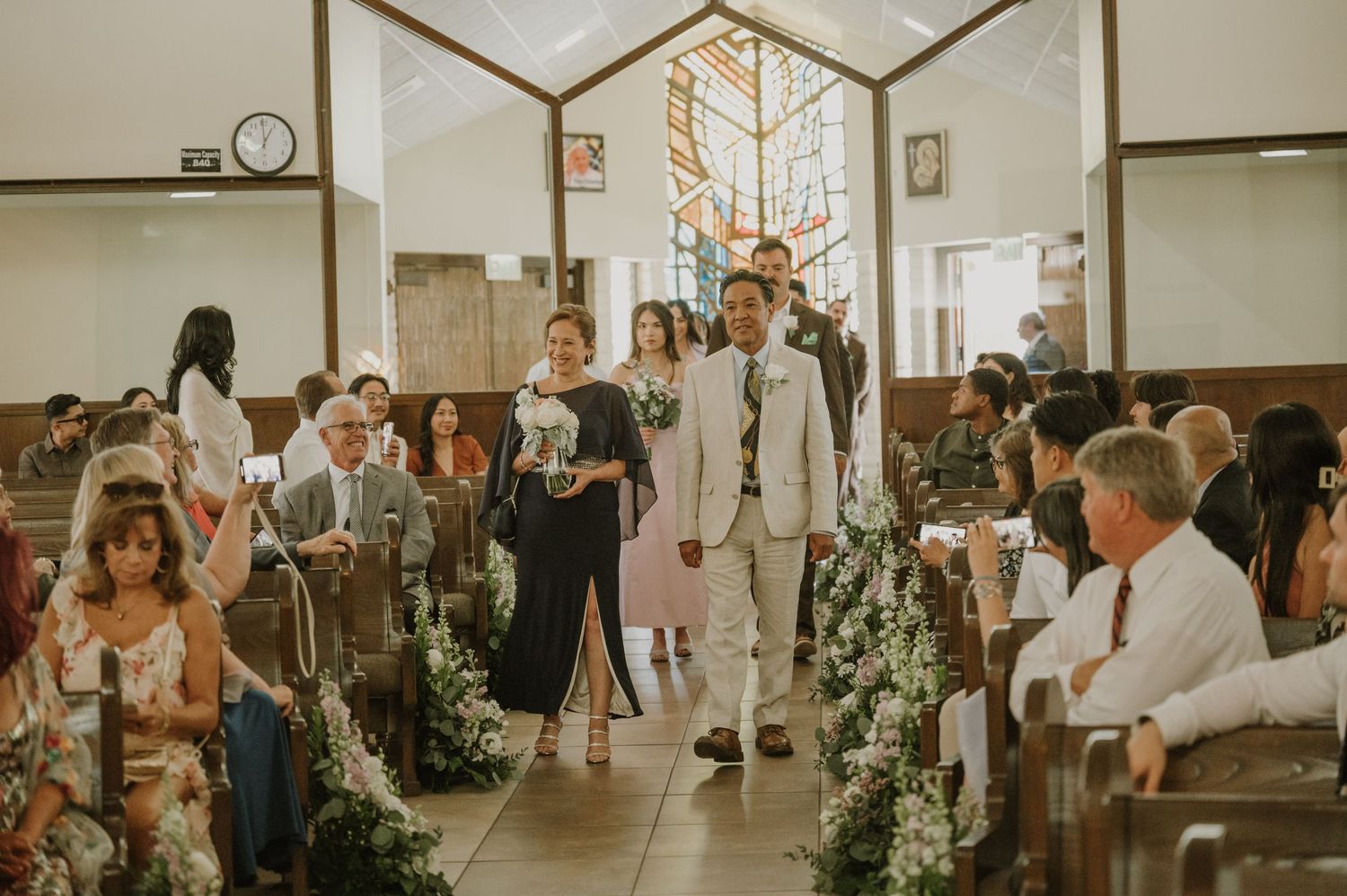 Groom walks down church aisle with family during wedding ceremony, flanked by floral arrangements and seated guests.