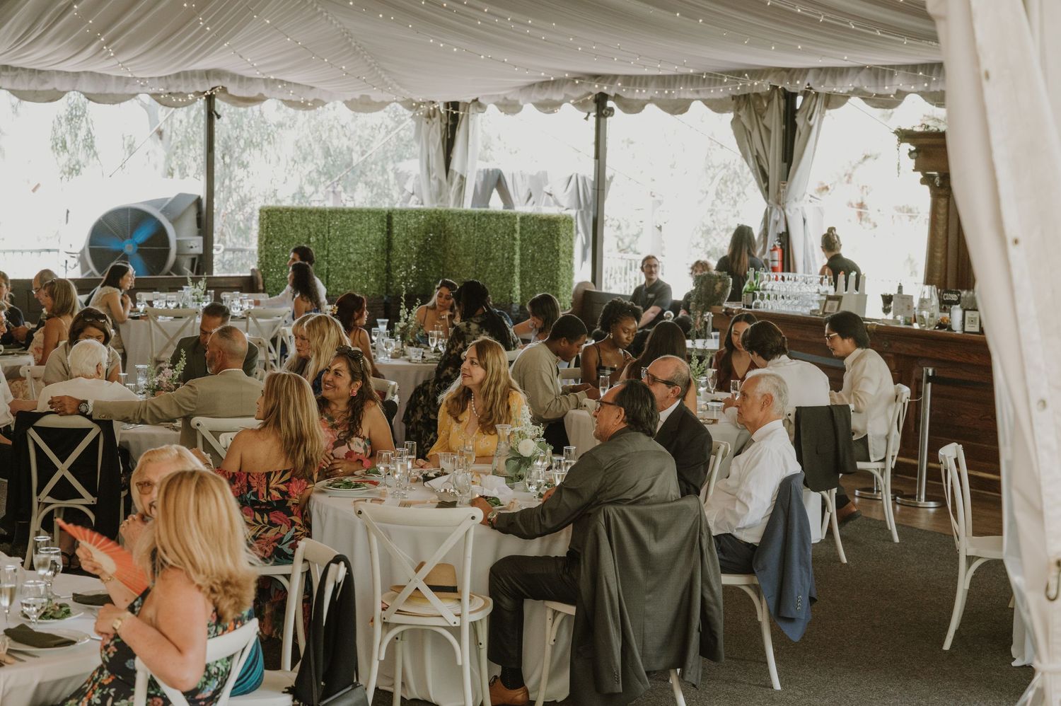 Guests dining under a white tent at an elegant outdoor wedding reception with round tables and white chairs.