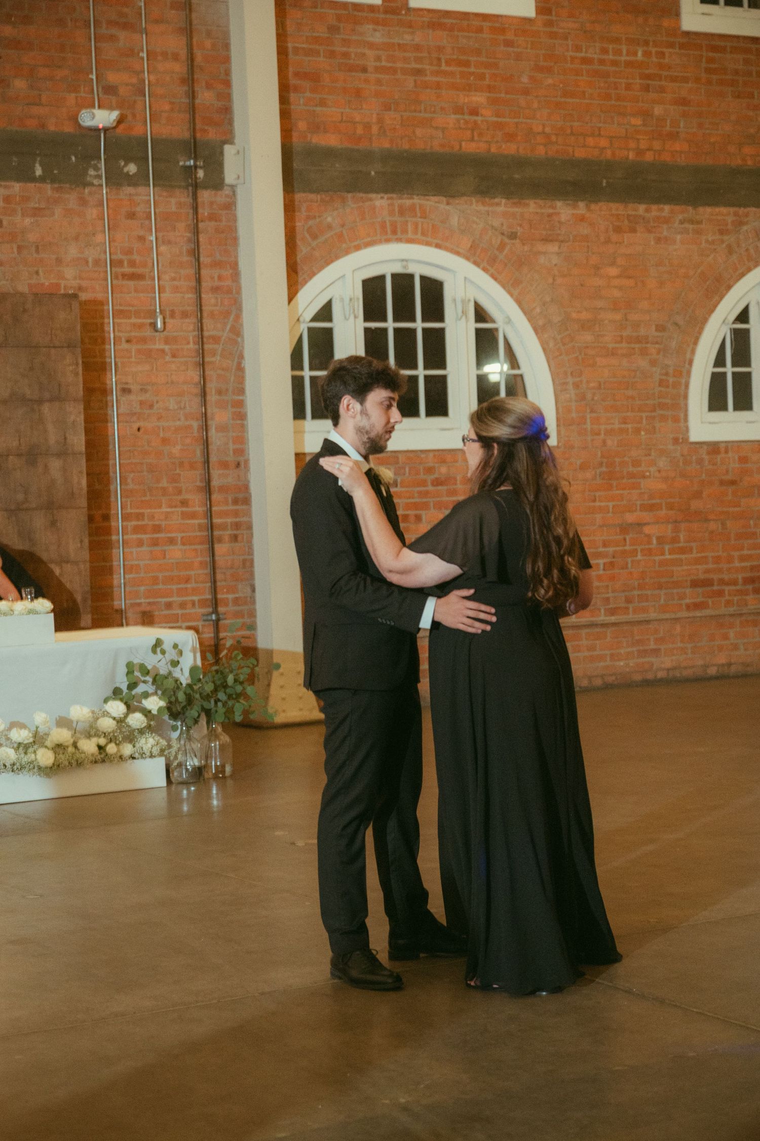 Groom shares a tender mother-son dance with woman in black gown at elegant brick wedding venue.
