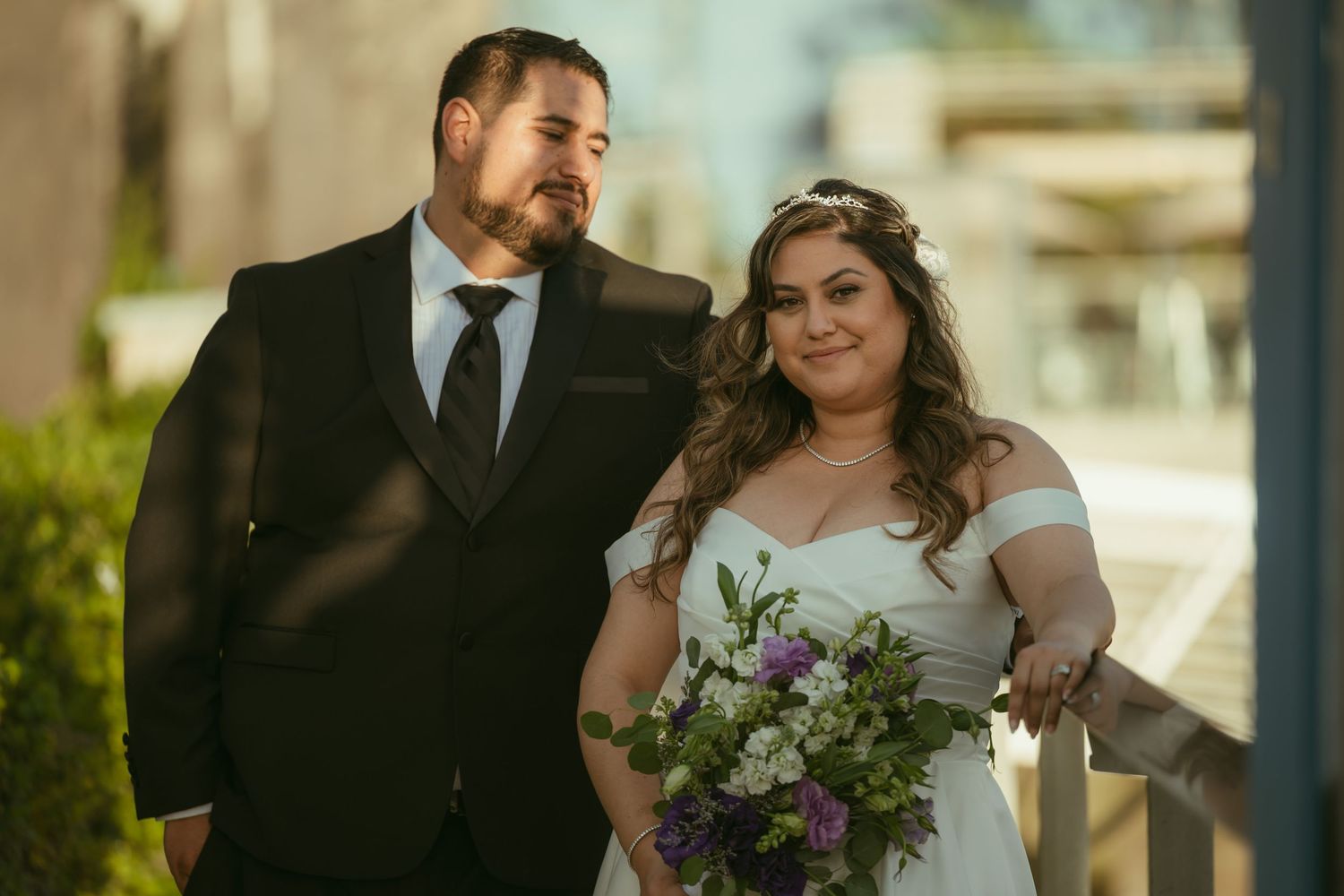 Bride and groom posing together outdoors, bride holding purple and white bouquet, groom in black suit with tie.