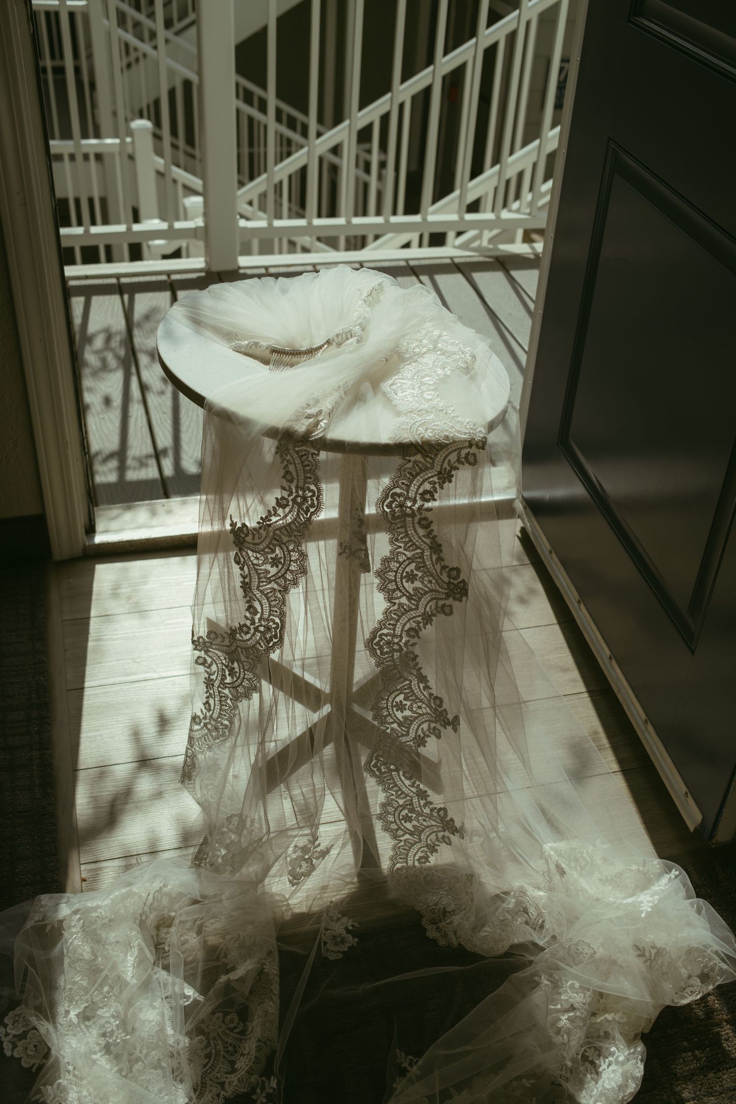 Elegant lace wedding veil draped over a wooden stool bathed in natural sunlight near a balcony door.
