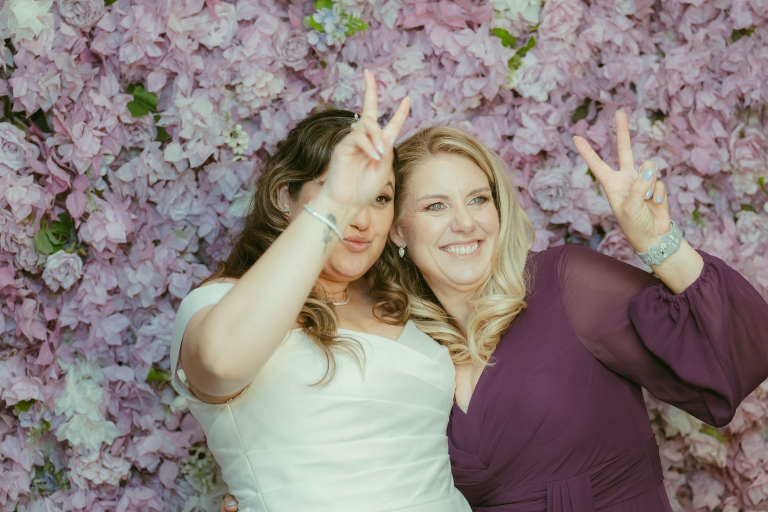 Bride and bridesmaid laugh and make peace signs lying on pink floral wall backdrop.