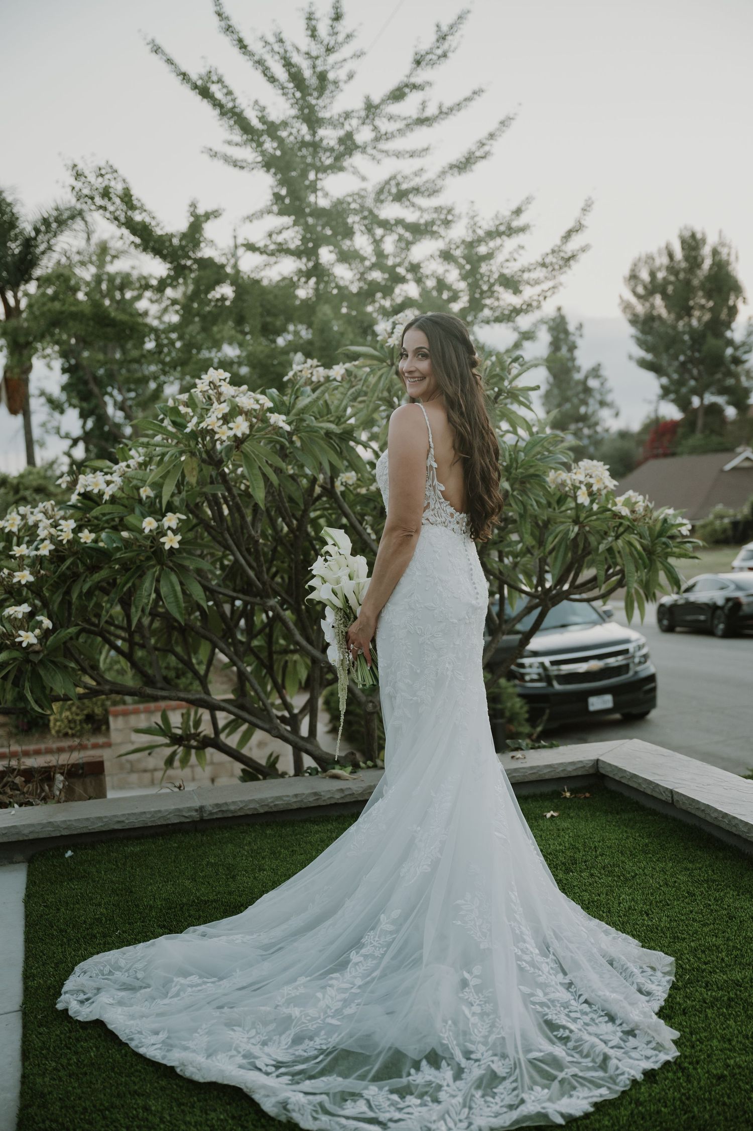 Bride in elegant lace mermaid wedding gown with long train poses outdoors near blooming white flowers.