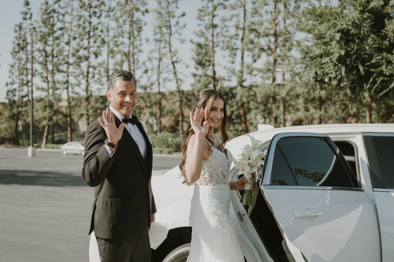 Bride and groom waving beside a white car on their wedding day, with trees in the background.