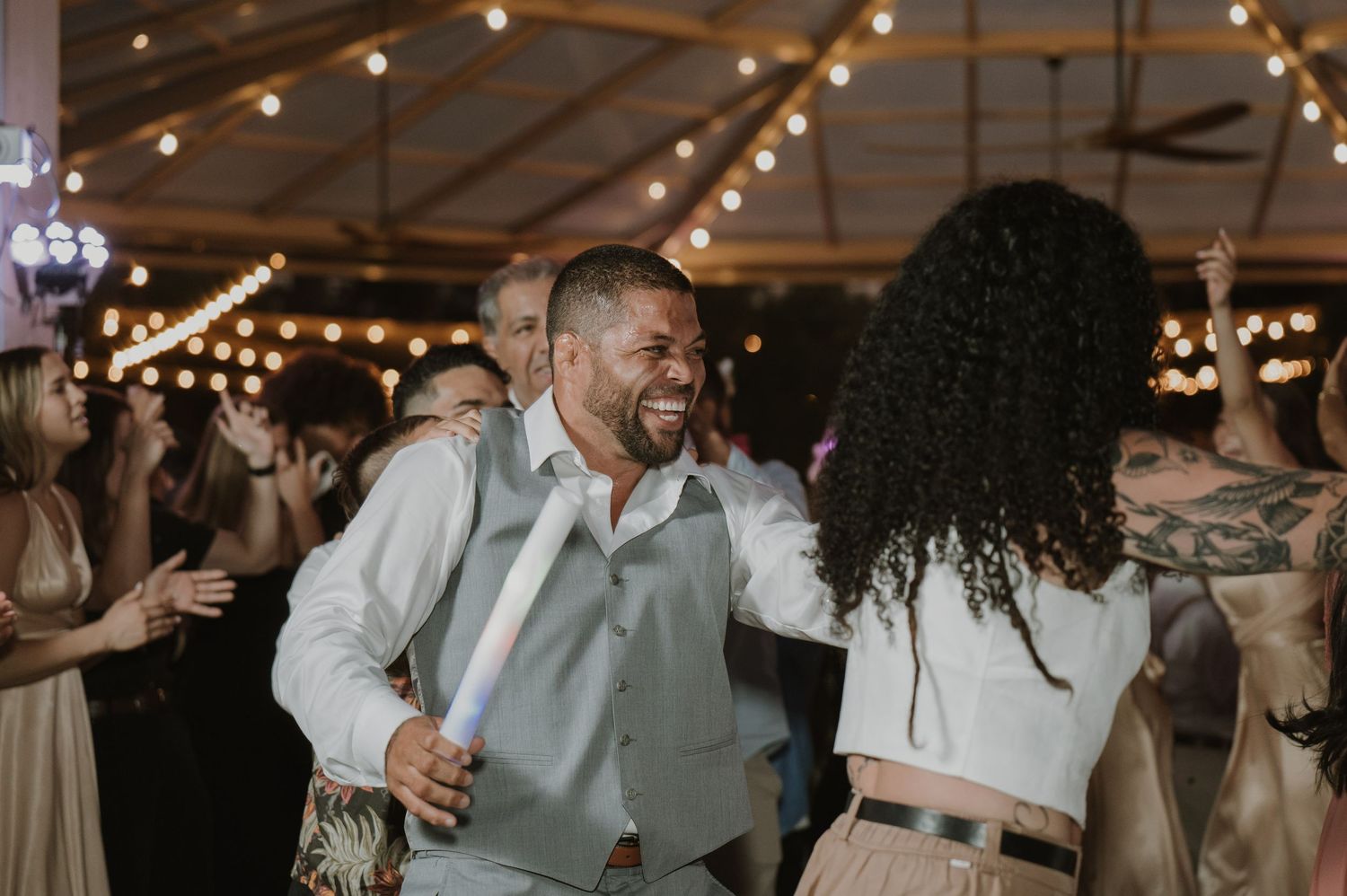 A smiling groom in a light blue vest dances with a guest under warm string lights at his wedding.