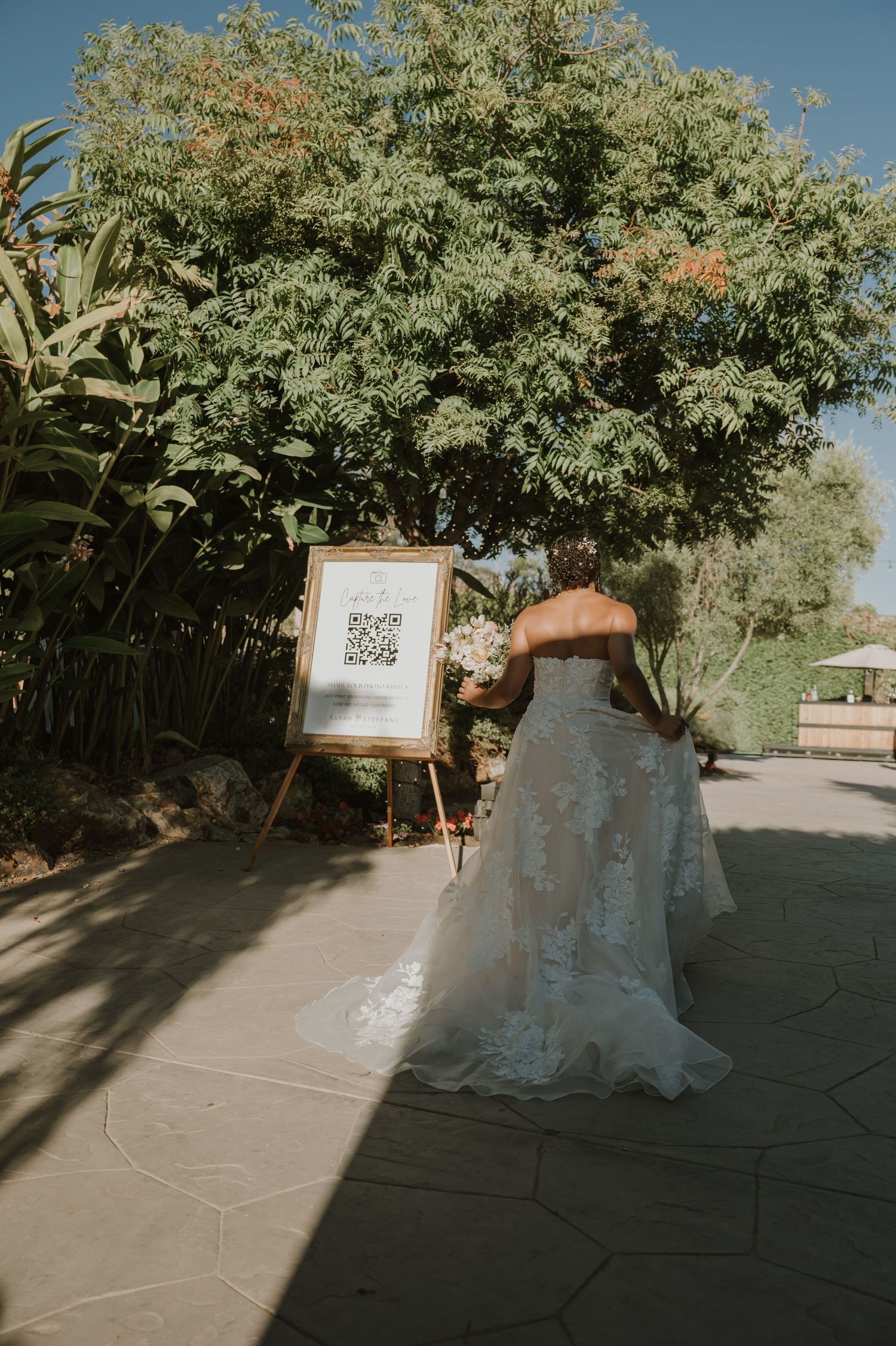 Bride in lace gown viewing QR code wedding sign in lush garden setting.
