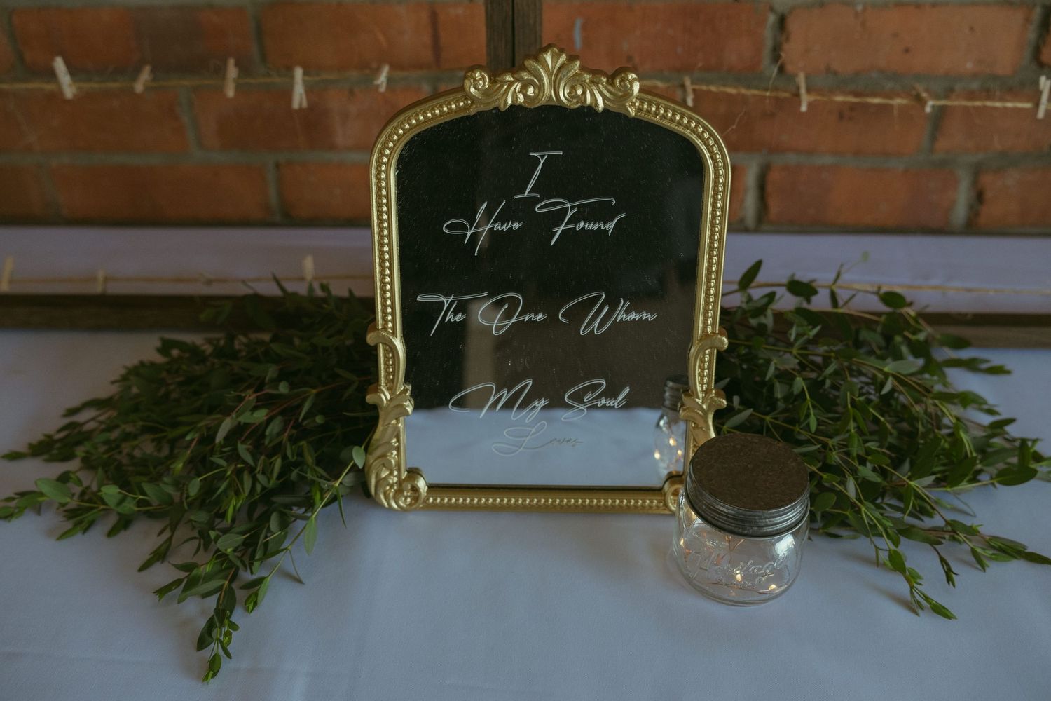 Gold ornate mirror with cursive wedding quote, surrounded by greenery and a mason jar on a white table.