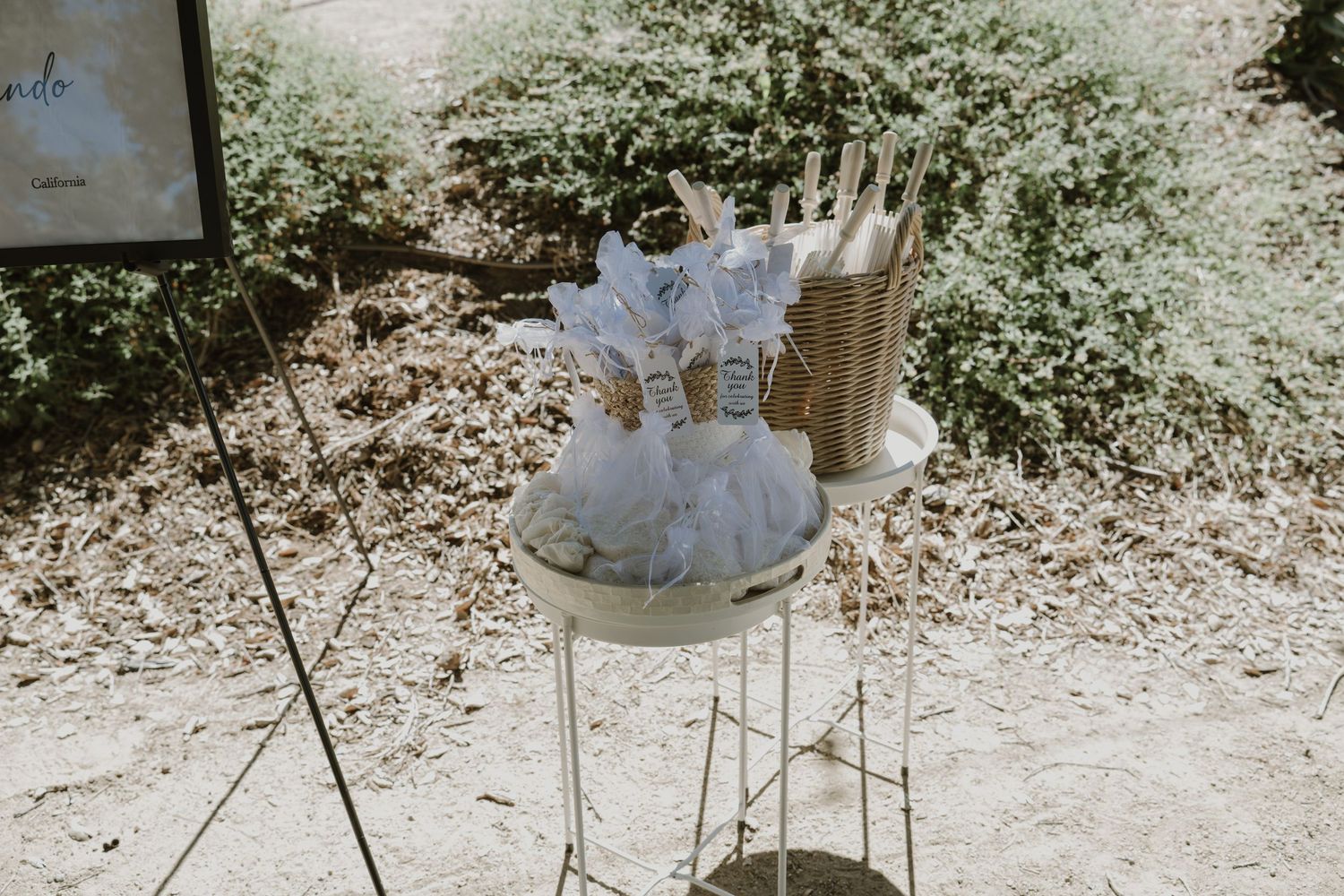 Outdoor wedding favor table with white tulle fans and wicker basket displayed on elegant stand in garden setting.