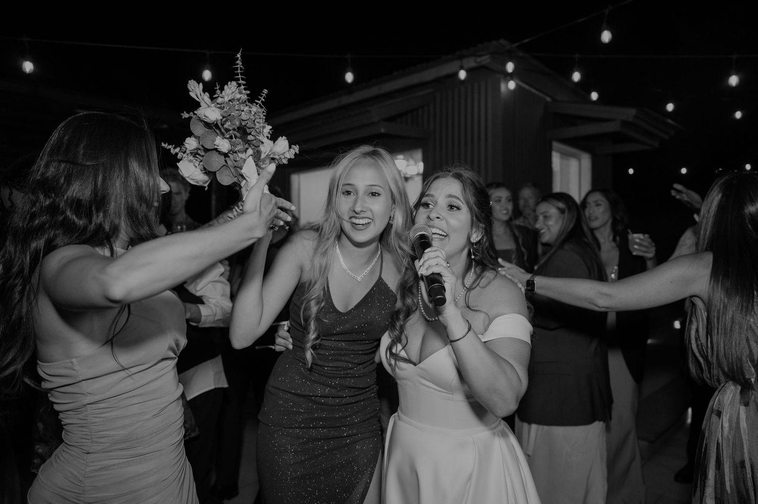Bride singing into microphone with bridesmaids holding bouquet aloft at joyful wedding reception dance floor.
