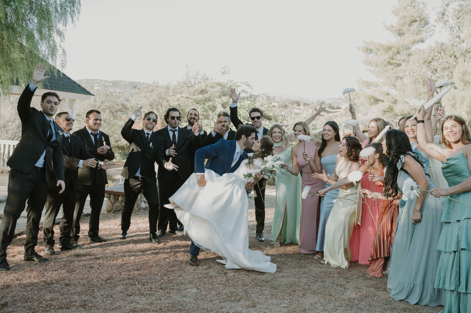 Excited wedding party cheers as groom lifts bride surrounded by bridesmaids and groomsmen outdoors.