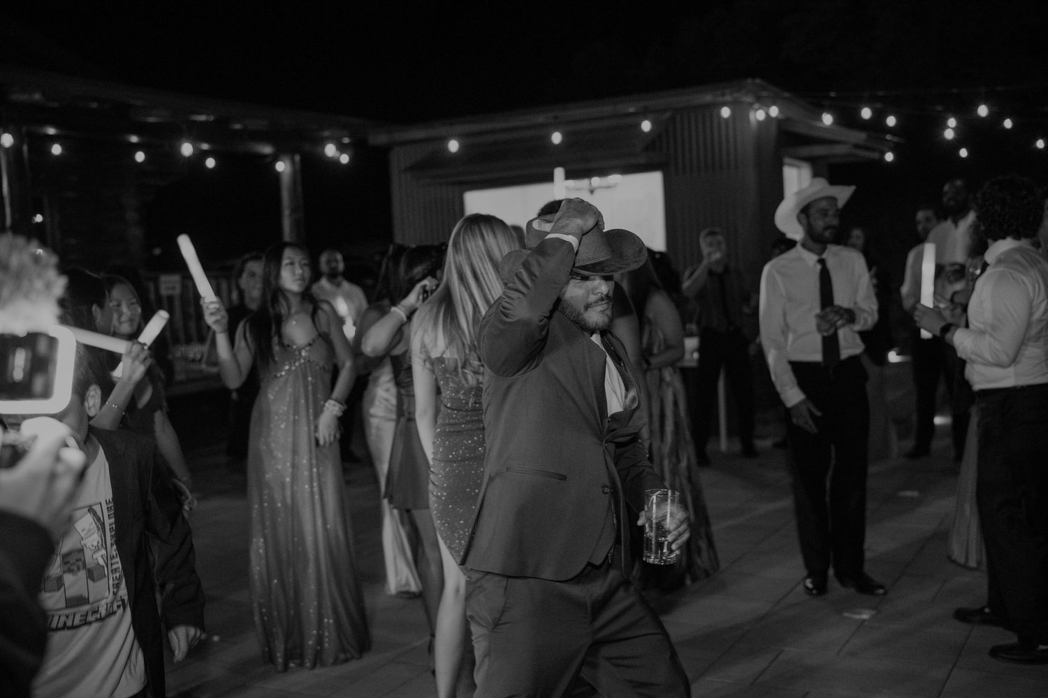 Black and white photo of wedding guests dancing under string lights at an outdoor evening reception.