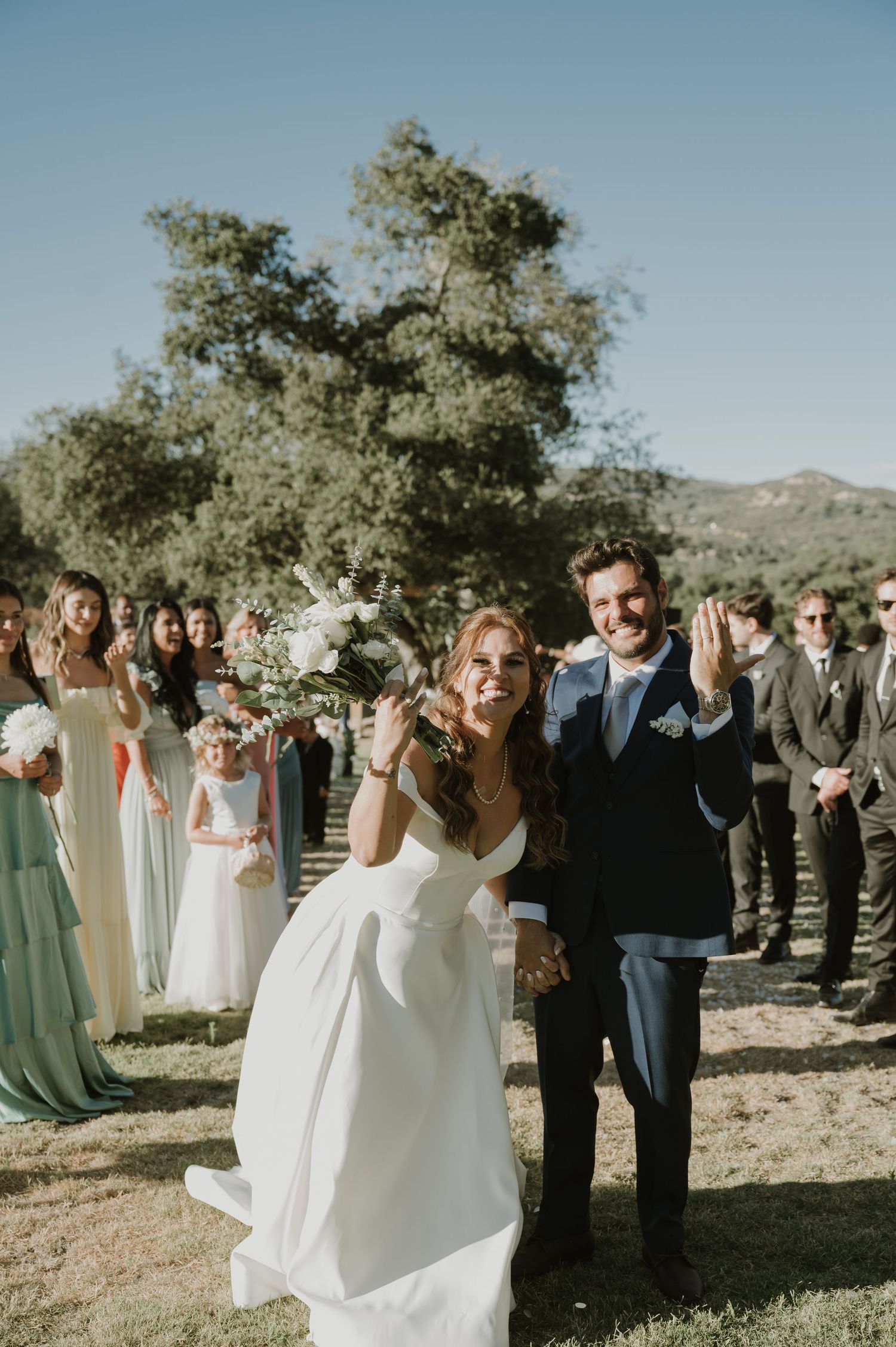 Joyful bride and groom exit outdoor wedding ceremony with bridesmaids in sage green dresses.
