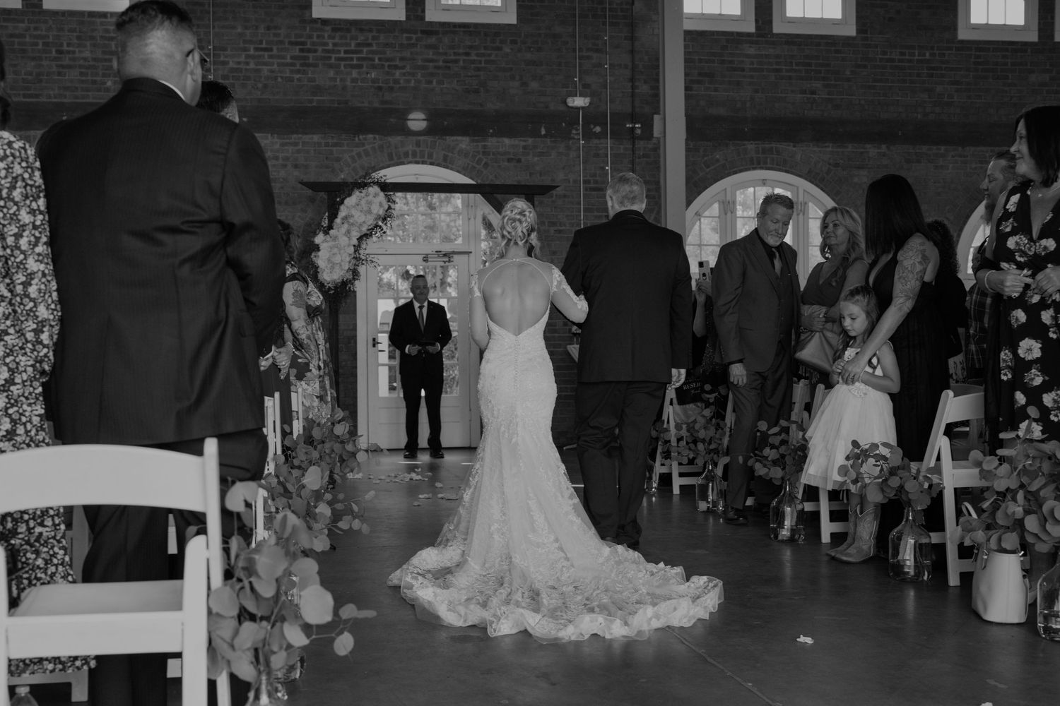 Black and white photo of a bride walking down the aisle in a lace gown at an elegant indoor wedding ceremony.