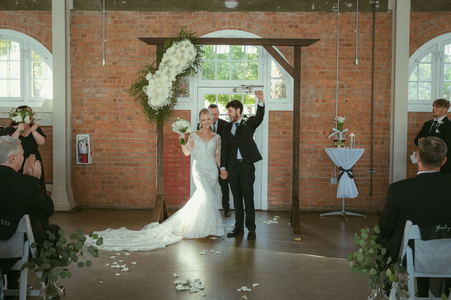Newlyweds celebrate with raised arms after wedding ceremony in rustic brick venue with floral arch.