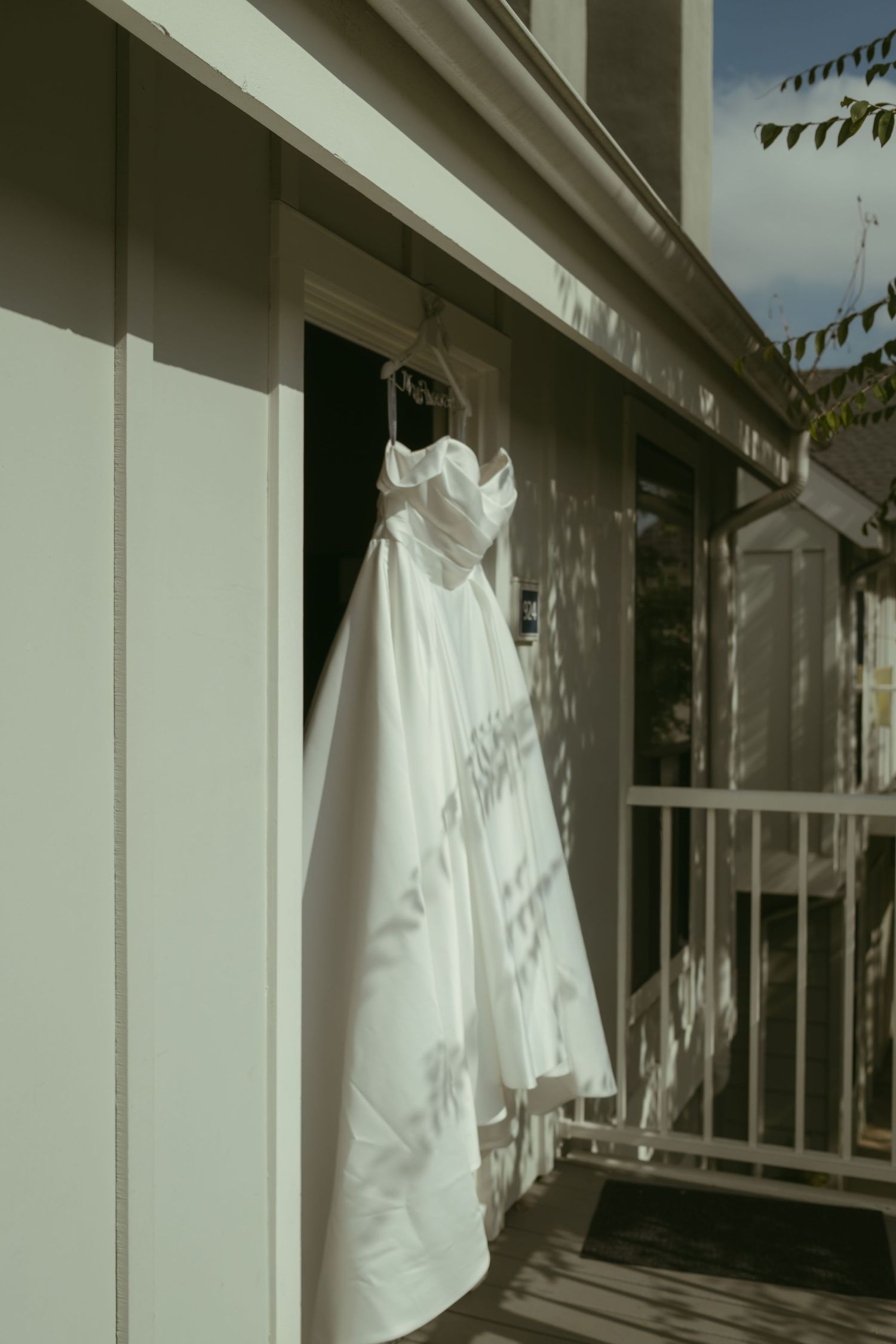 White wedding dress hanging on a door frame outdoors, with sunlight casting leaf shadows on the satin fabric.
