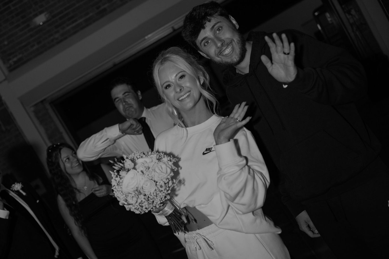 Black and white photo of smiling bride holding bouquet with groom waving outside.
